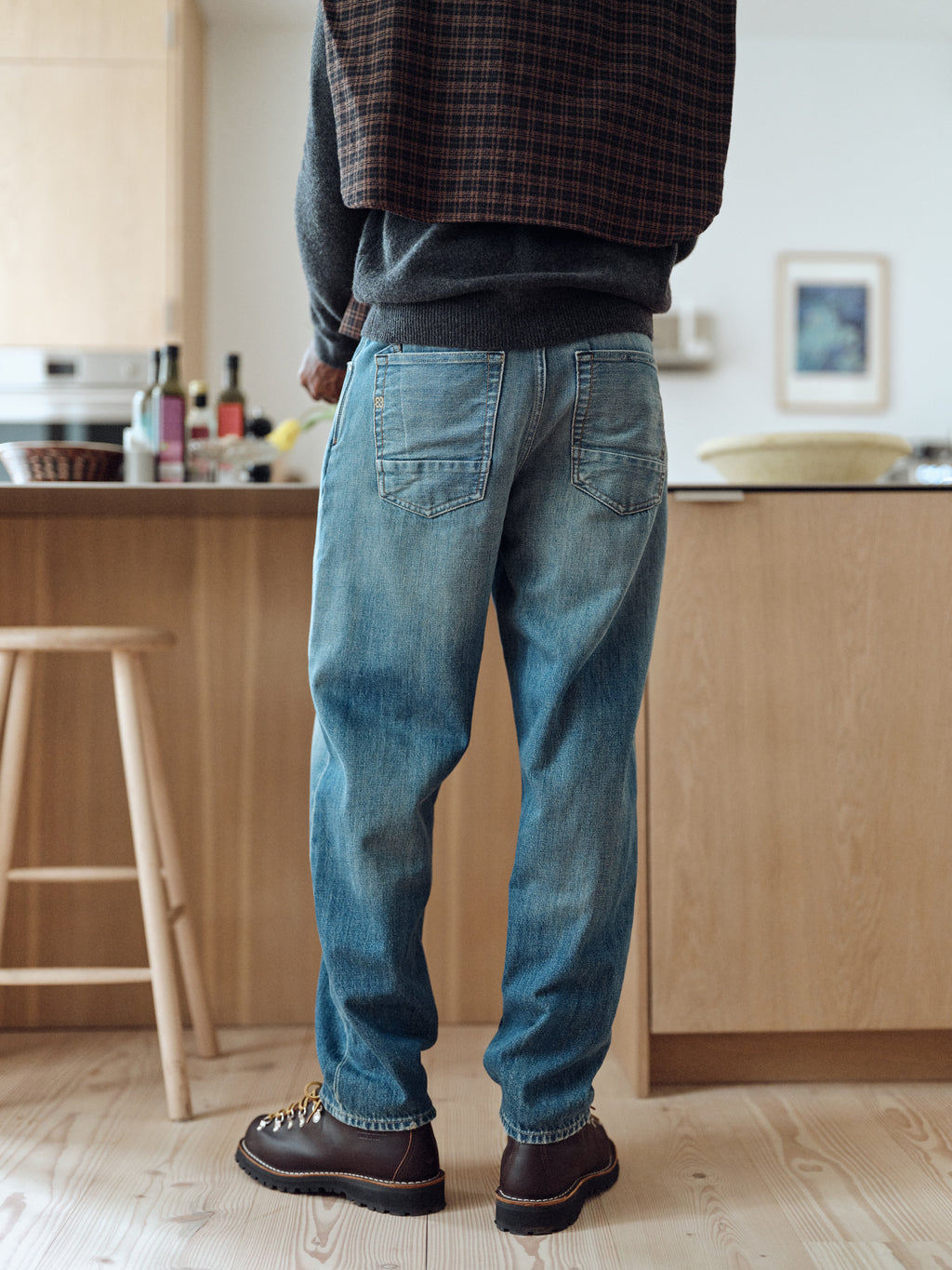 A person wearing Gabba GABsenna Wolv DS0004 Jeans in dark blue denim and dark shoes stands in a kitchen with their back to the camera, a plaid jacket draped over their shoulders. Bottles and bowls are visible on the countertop.