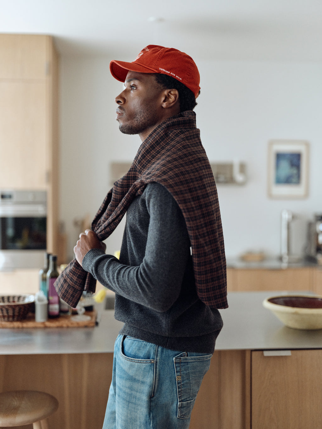 A man in a red cap and jeans stands in a modern kitchen, with a plaid shirt draped over his shoulders and the Gabba GABworren Polo Aico LS Knit in Dark Grey Mel layered beneath, its ribbed polo collar adding timeless style as he looks thoughtfully into the distance.