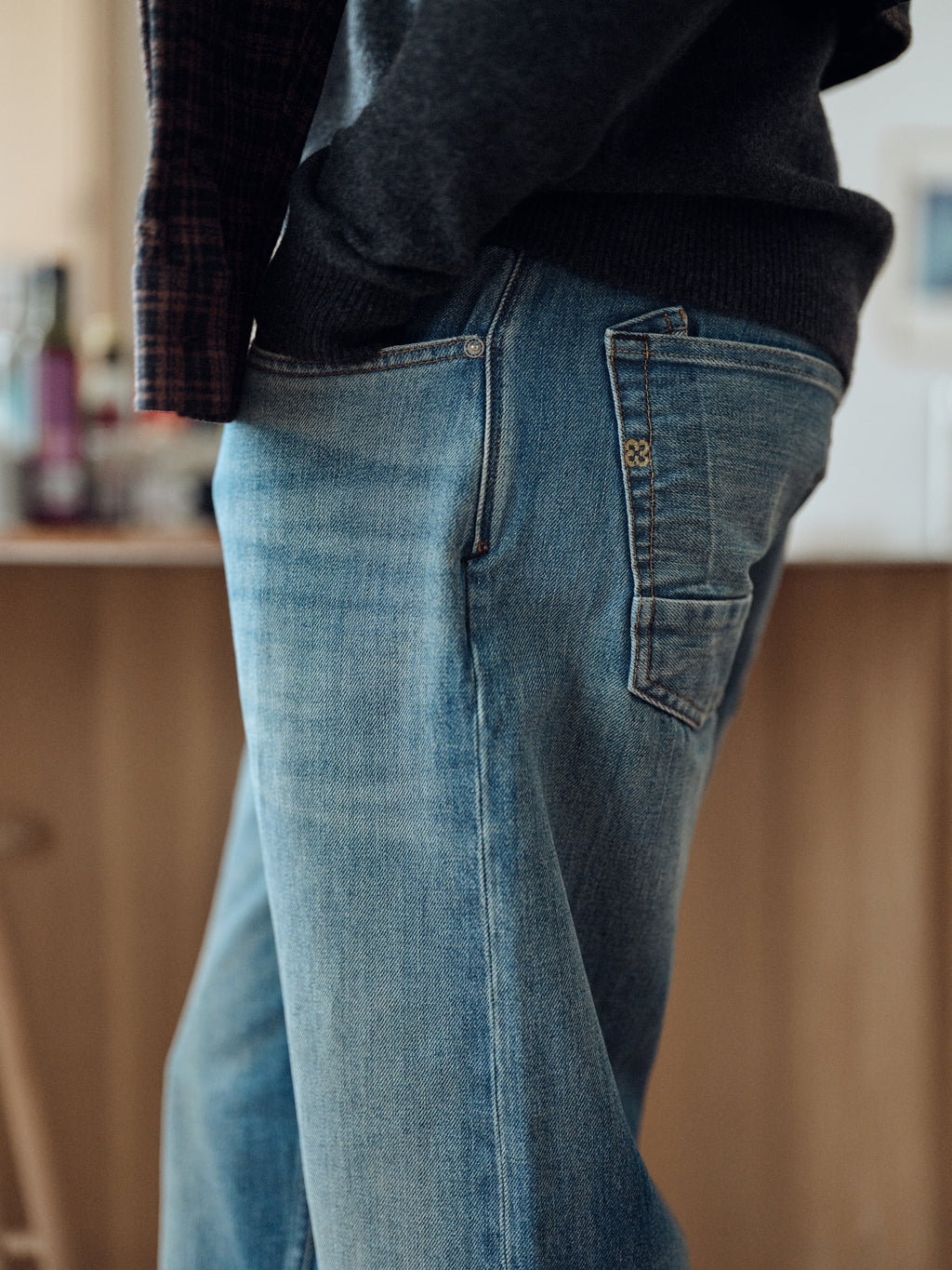A person wearing Gabba’s GABsenna Wolv DS0004 Jeans in dark blue denim and a dark sweater stands indoors with one hand in their pocket, facing away from the camera. The softly blurred background reveals household items and wooden furniture.