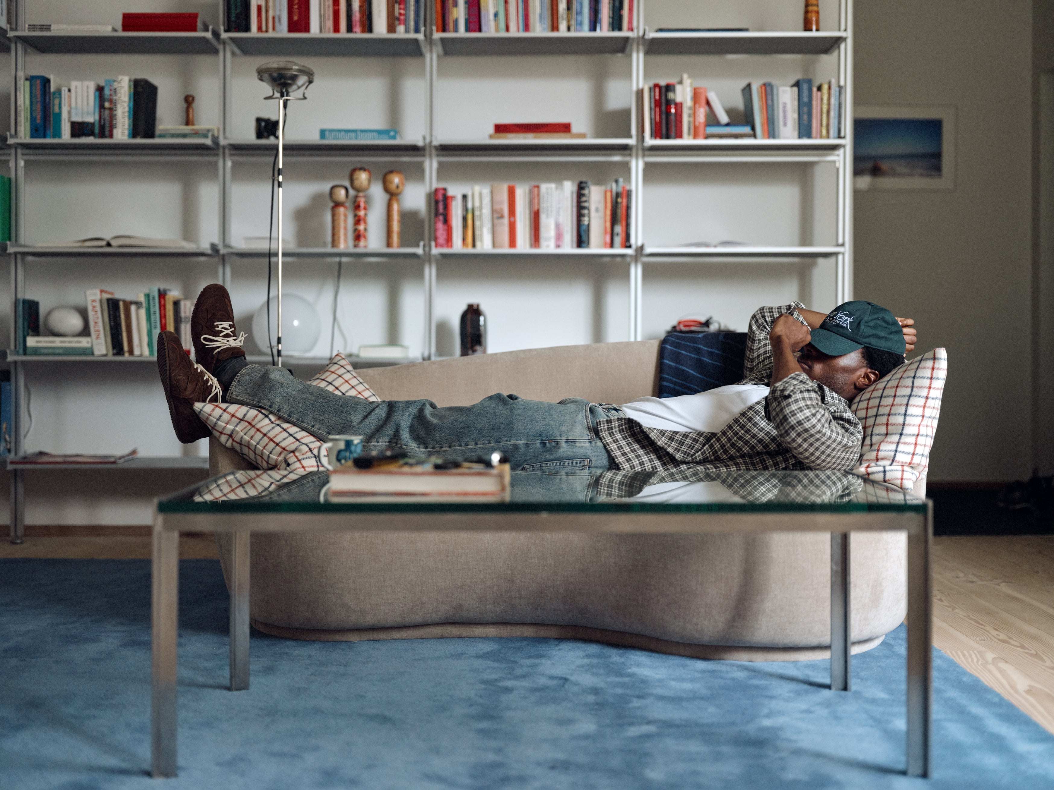 Wearing Gabba’s GABsven Reg TX2 LS Shirt in Forest Night—a yarn-dyed cotton, regular-fit shirt—paired with jeans, a checked jacket, and a green cap, the person is lying on a sofa in a living room featuring bookshelves, a blue rug, and a glass coffee table.