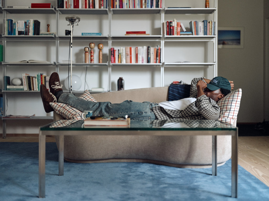 Wearing Gabba’s GABsven Reg TX2 LS Shirt in Forest Night—a yarn-dyed cotton, regular-fit shirt—paired with jeans, a checked jacket, and a green cap, the person is lying on a sofa in a living room featuring bookshelves, a blue rug, and a glass coffee table.