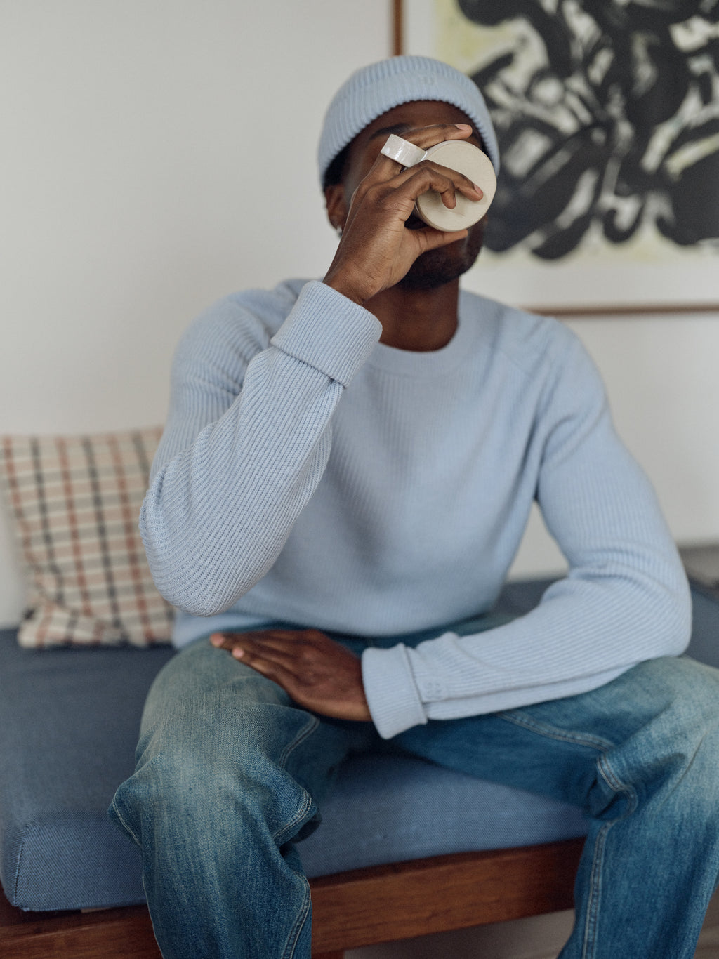 A person in the Gabba GABremy O-Neck Flash LS Knit in Dusty Blue and a beanie sits on a bench, sipping from a mug. A plaid pillow and abstract artwork can be seen in the background.