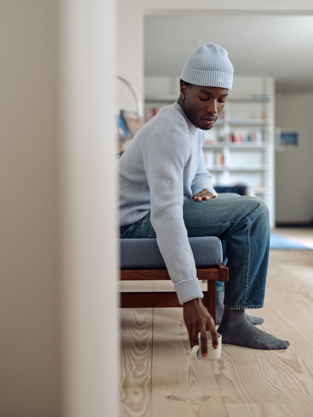 Wearing the Gabba GABremy O-Neck Flash LS Knit in Dusty Blue, a person in jeans and a beanie sits on a bench and reaches down to pick up a white mug from the wooden floor of a modern, bright room.