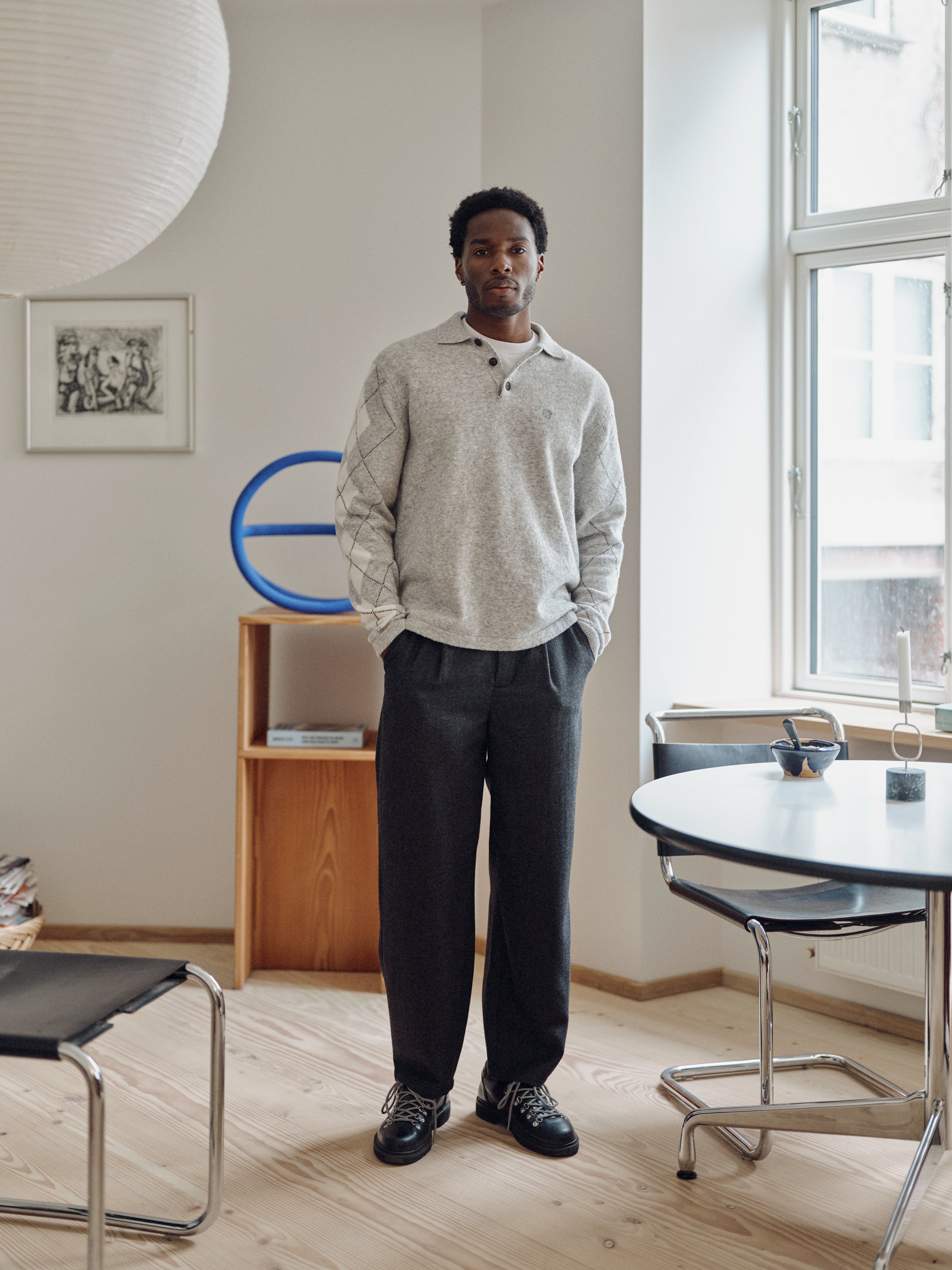 Wearing the Gabba GABdavid Twilight Pant in Dark Grey, a man stands in a modern, minimal living space with light wood floors, a round table, chairs, and large windows allowing natural light to fill the room.