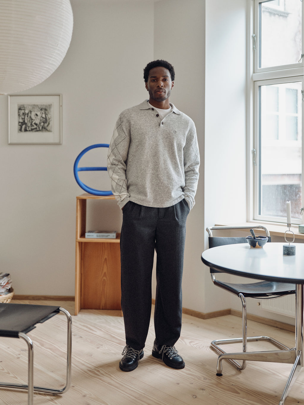 Wearing the Gabba GABdavid Twilight Pant in Dark Grey, a man stands in a modern, minimal living space with light wood floors, a round table, chairs, and large windows allowing natural light to fill the room.