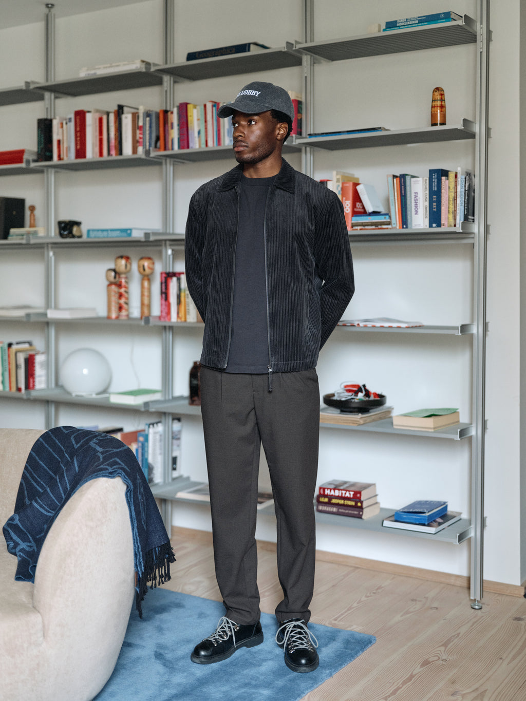 A man wearing the Gabba GABnik Zip Riglia GOTS Jacket in dark grey—modern outerwear crafted from organic cotton corduroy—stands indoors by shelves filled with books and decor. Dressed in dark clothing and a cap, he looks to his left with his hands behind his back, next to a beige chair draped with a dark blue blanket.