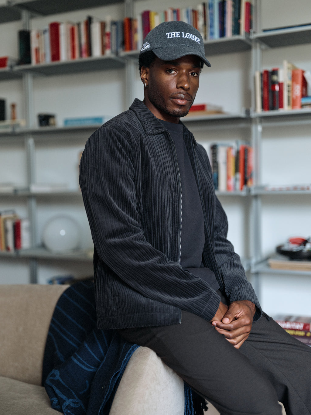 A man in a contemporary outfit—the Gabba GABnik Zip Riglia GOTS Jacket in dark grey, crafted from organic cotton corduroy—sits on a beige couch in a modern room with bookshelves filled with books in the background.
