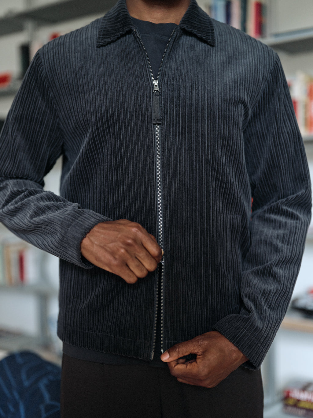 A person wearing the Gabba GABnik Zip Riglia GOTS Jacket in Dark Grey stands indoors, holding the zipper, with shelves of books visible in the blurred background.
