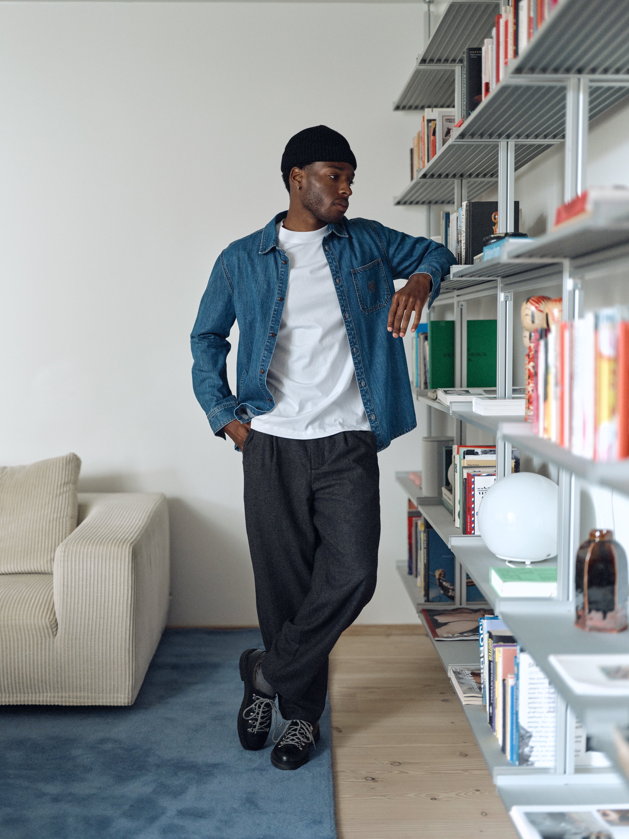 A man in a denim shirt, white t-shirt, and Gabba GABdavid Twilight Pant - Dark Grey leans on a bookshelf in a modern living room, looking to the side.