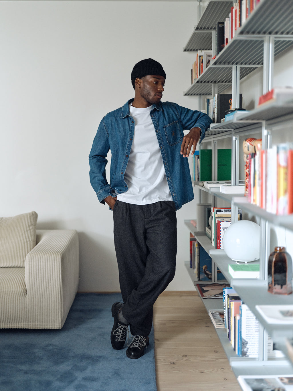 A man in a denim shirt, white t-shirt, and Gabba GABdavid Twilight Pant - Dark Grey leans on a bookshelf in a modern living room, looking to the side.
