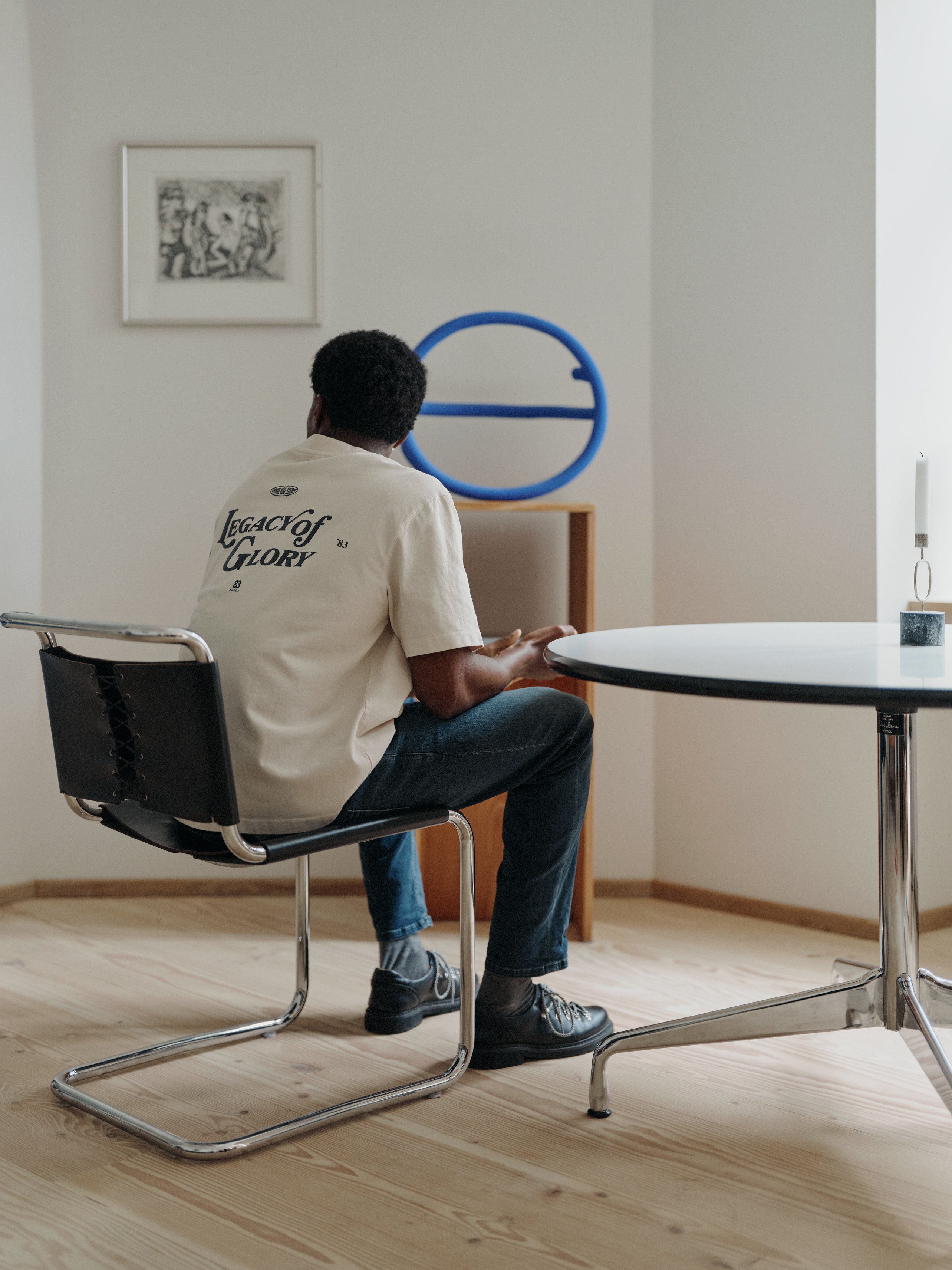 A person sits on a black chair at a round table, facing away, wearing the Gabba GABlegin Boxy Glory SS T-Shirt in Birch—featuring a graphic back print and cut in a relaxed, boxy fit textured cotton jersey. A blue circular sculpture and framed artwork decorate the minimal, well-lit room with wooden floors.