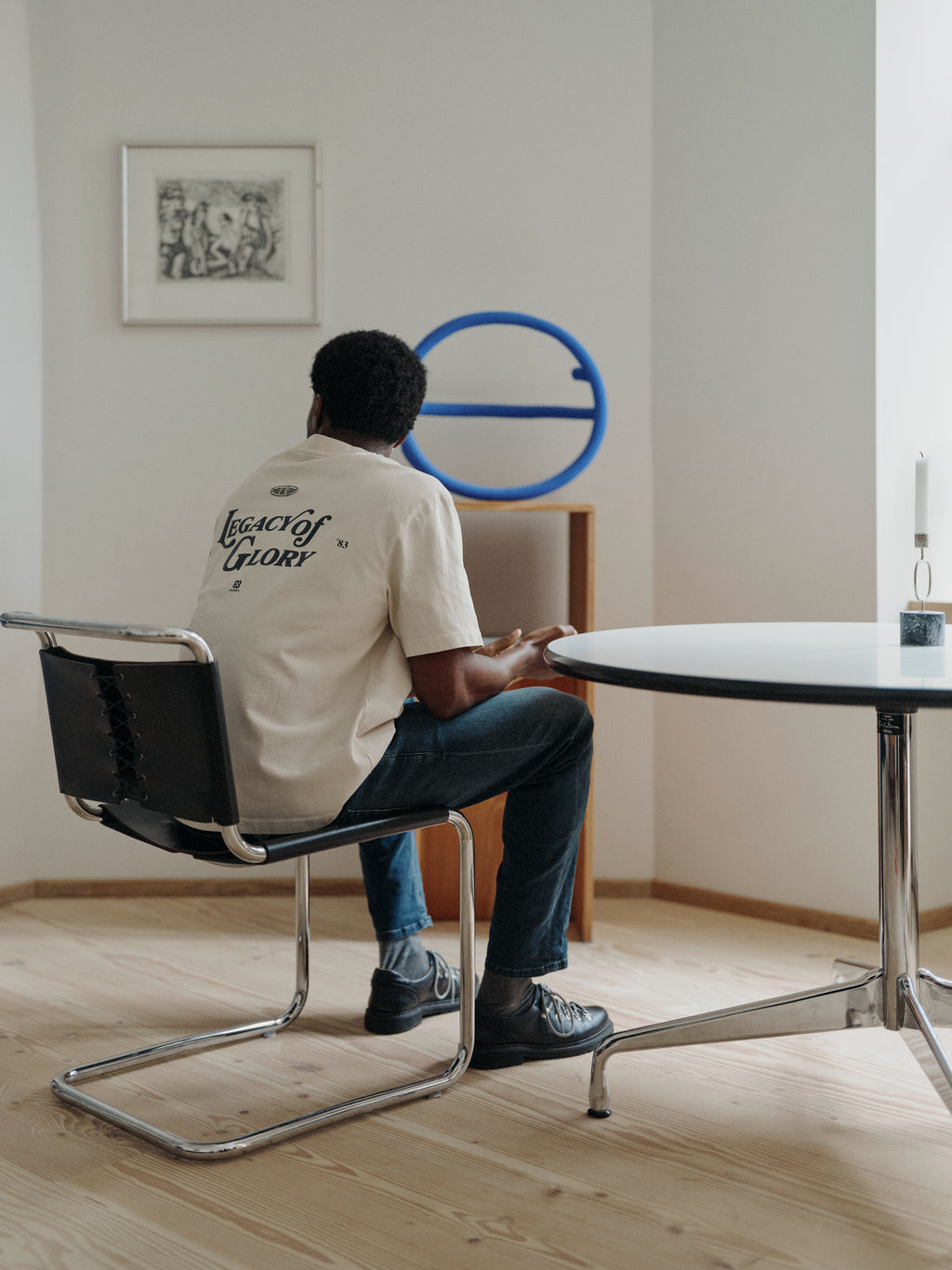 A person sits on a black chair at a round table, facing away, wearing the Gabba GABlegin Boxy Glory SS T-Shirt in Birch—featuring a graphic back print and cut in a relaxed, boxy fit textured cotton jersey. A blue circular sculpture and framed artwork decorate the minimal, well-lit room with wooden floors.
