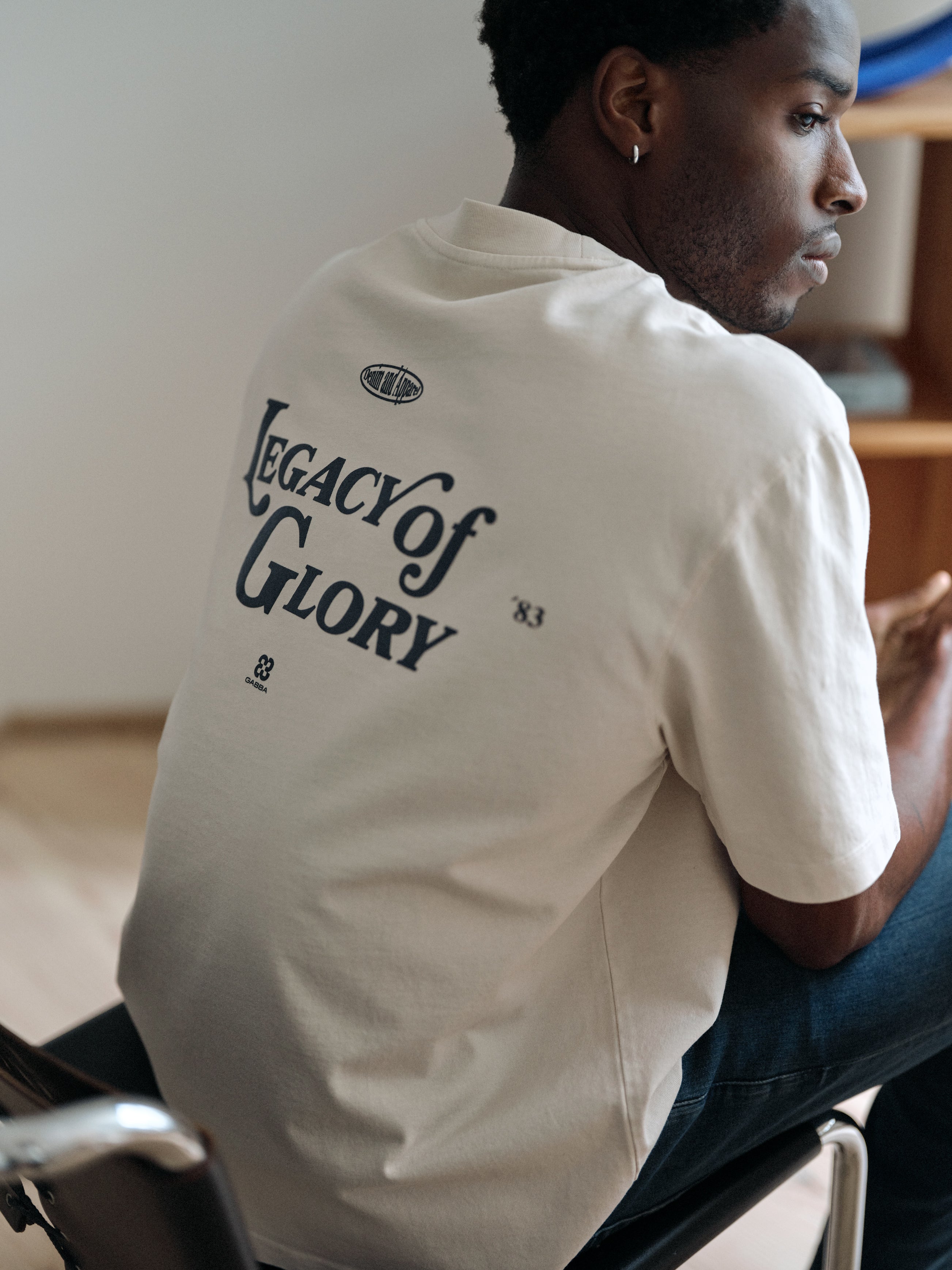 A man sits in a chair, facing sideways. He wears the Gabba GABlegin Boxy Glory SS T-Shirt in Birch—a light-colored, textured cotton jersey tee featuring “Legacy of Glory” boldly printed on the back as a striking graphic detail.