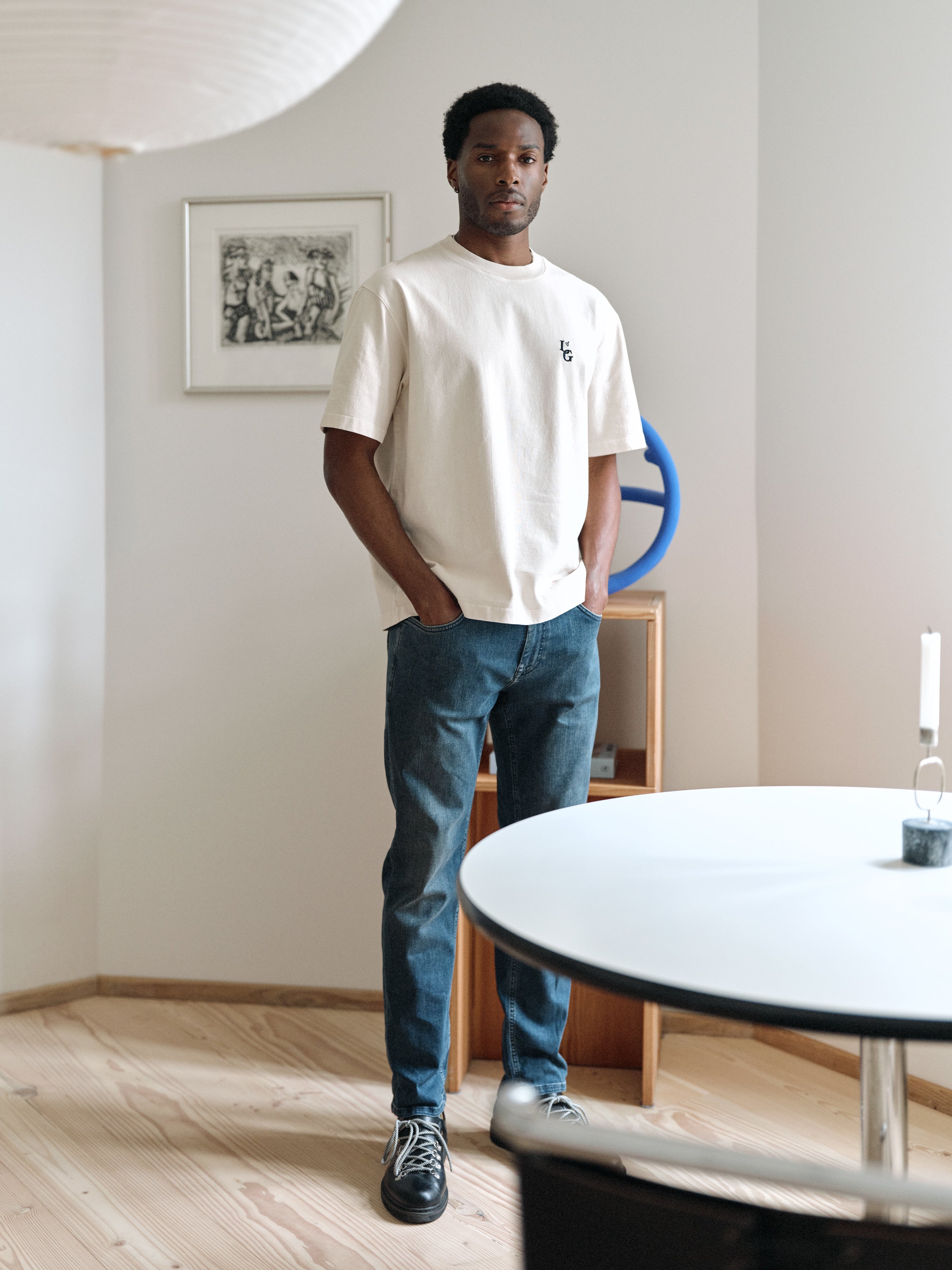 A man wearing the Gabba GABlegin Boxy Glory SS T-Shirt in Birch, featuring a graphic back print and relaxed boxy fit in textured cotton jersey, pairs it with jeans as he stands in a modern, minimalist room with light wood floors, a round table, candle, wall art, and a blue sculpture—an ensemble that perfectly complements the contemporary ambiance.