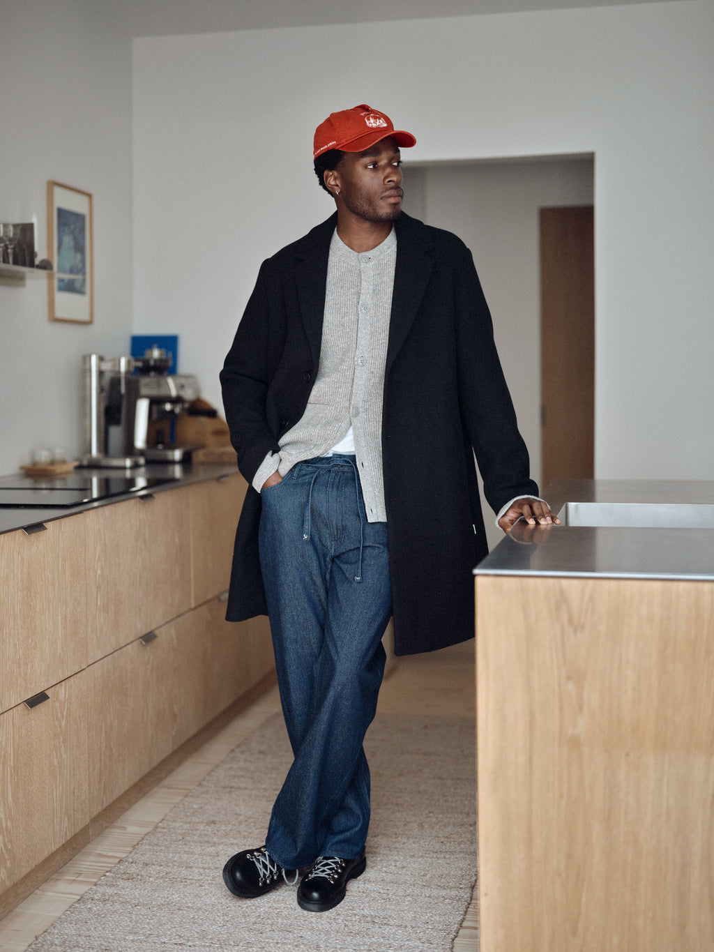 A person wearing a red cap, black coat, gray sweater, and Gabba GABnoah Tona Rinse Jeans in dark blue denim stands in a modern kitchen with light wood cabinets, looking to the side with one hand in their pocket. Black shoes complete the casual yet refined look.