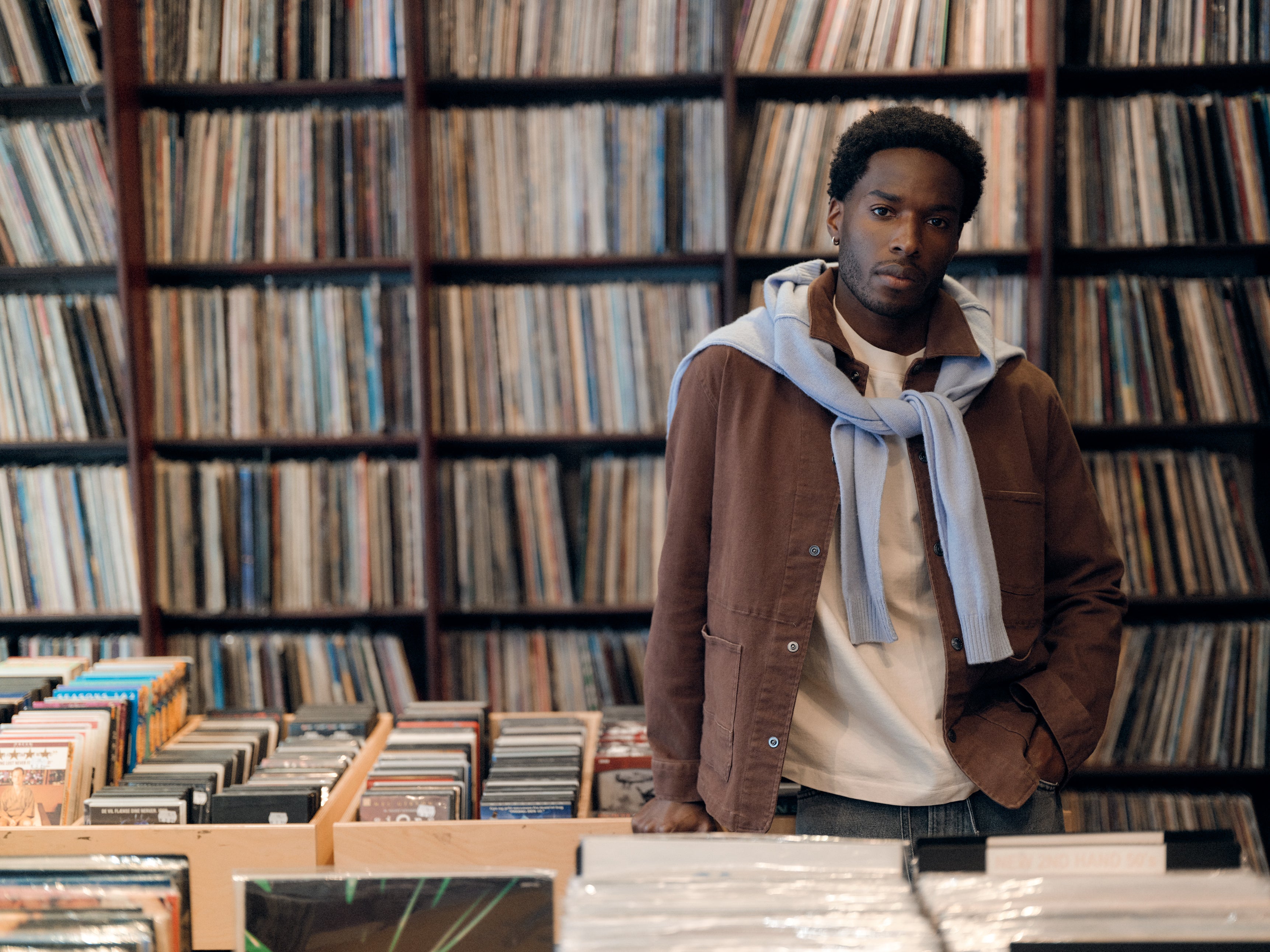 A man stands in a record store, surrounded by shelves filled with vinyl records. He wears a light blue sweater draped over a brown jacket and white shirt, complemented by Gabba’s GABbeta Twill - Brown Slate regular fit garment-dyed stretch twill pants with utility pockets, looking thoughtfully at the camera.