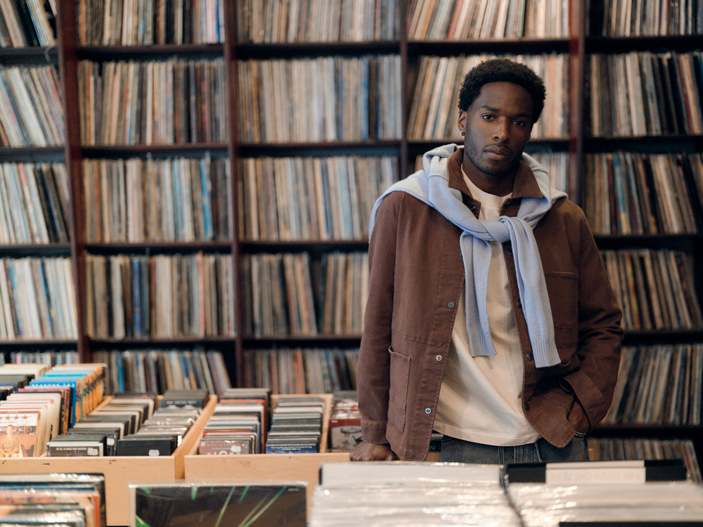 A man stands in a record store, surrounded by shelves filled with vinyl records. He wears a light blue sweater draped over a brown jacket and white shirt, complemented by Gabba’s GABbeta Twill - Brown Slate regular fit garment-dyed stretch twill pants with utility pockets, looking thoughtfully at the camera.