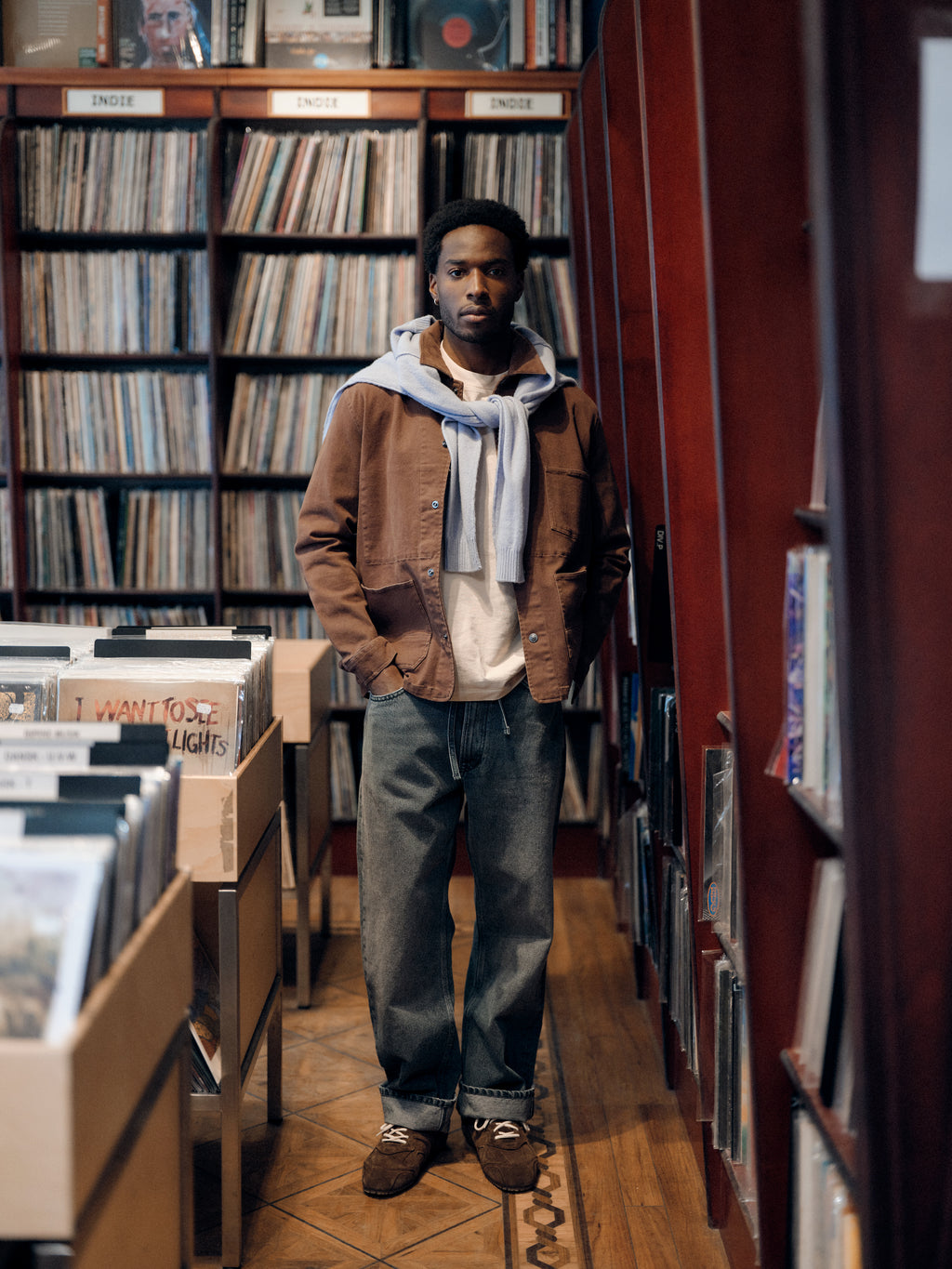 A person stands in a record store, surrounded by shelves of vinyl records. They wear a brown jacket, white shirt, sweater draped over their shoulders, and Gabba GABnoah Primesto WW0030 Jeans in Grey Denim paired with brown shoes, looking at the camera with a neutral expression.