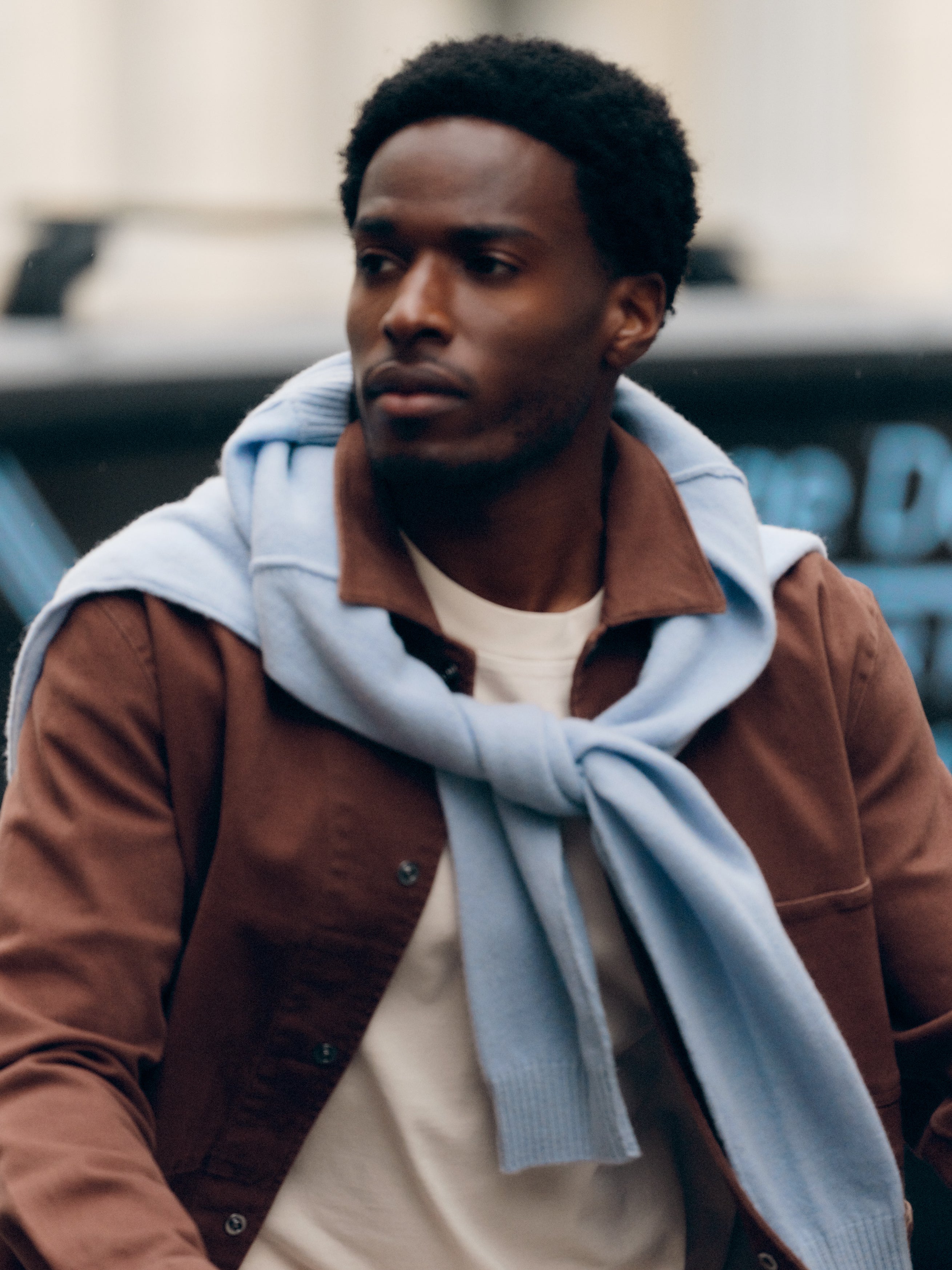 A man wearing the Gabba GABbeta Twill jacket in Brown Slate with a light blue sweater draped over his shoulders walks outdoors, glancing to the side—showcasing a versatile layering essential perfect for any season.
