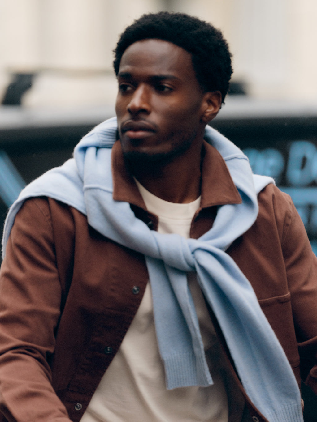 A man wearing the Gabba GABbeta Twill jacket in Brown Slate with a light blue sweater draped over his shoulders walks outdoors, glancing to the side—showcasing a versatile layering essential perfect for any season.