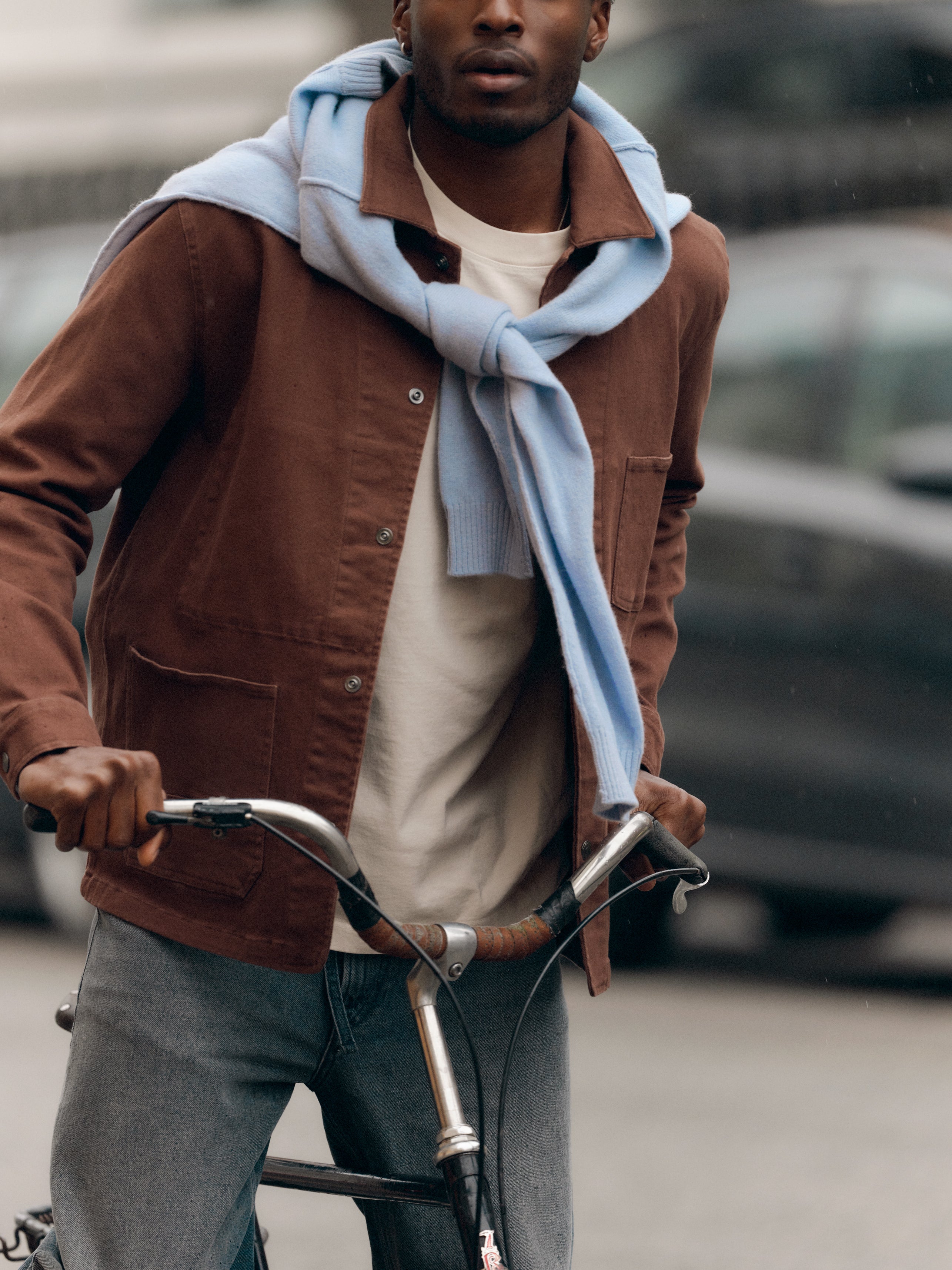 A person wearing the Gabba GABlegin Boxy Glory SS T-Shirt in Birch, layered under a brown jacket and draped with a textured light blue cotton jersey over their shoulders, stands outdoors holding a bicycle handlebar, with cars blurred in the background.