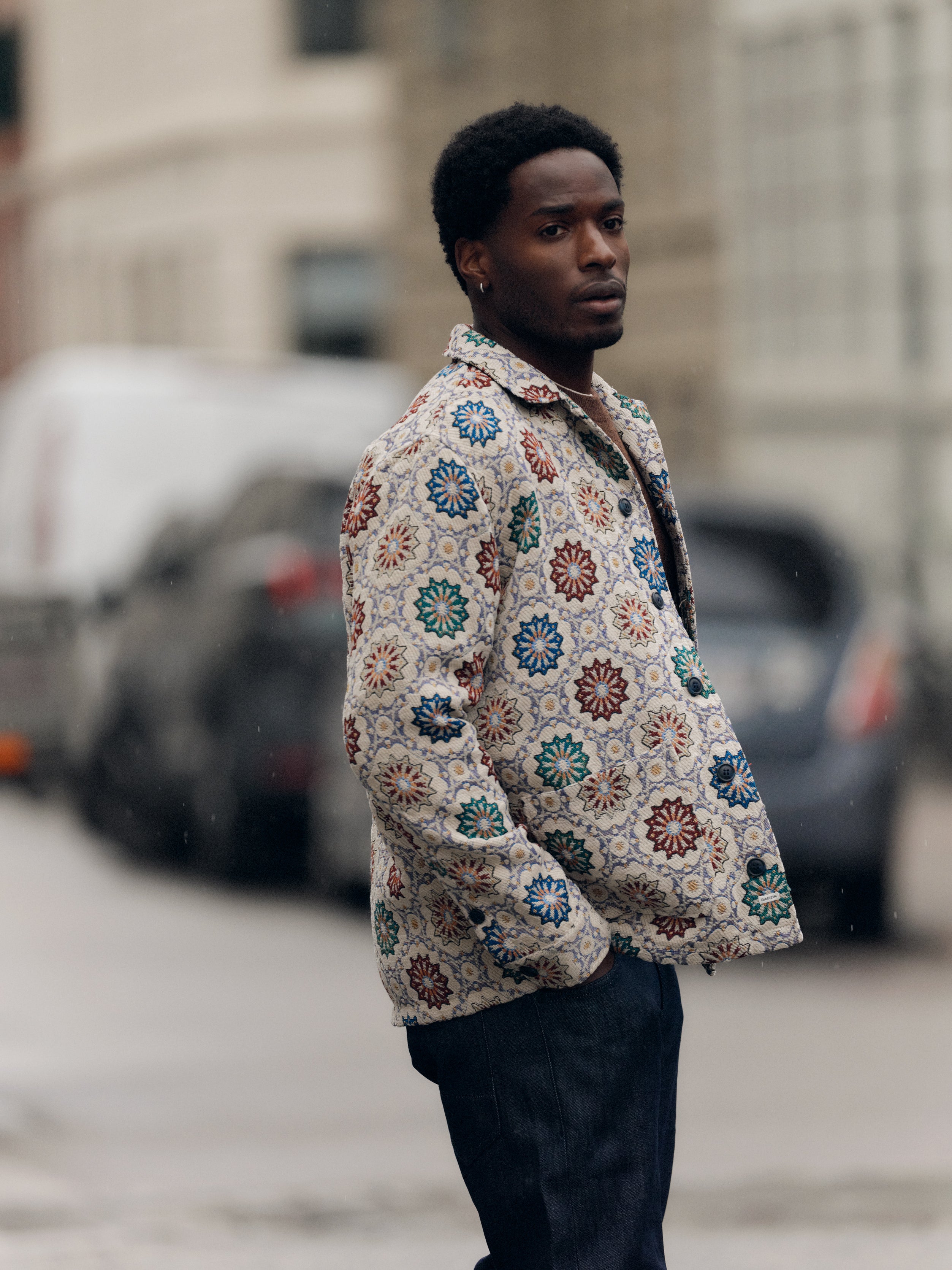 A man stands outdoors on a city street, wearing the Gabba GABsimone Nava LS - Birch jacket, which features a resort collar and colorful floral designs in textured jacquard fabric. He pairs it with dark jeans and looks slightly to the side with a neutral expression. Blurred cars and buildings are visible in the background.
