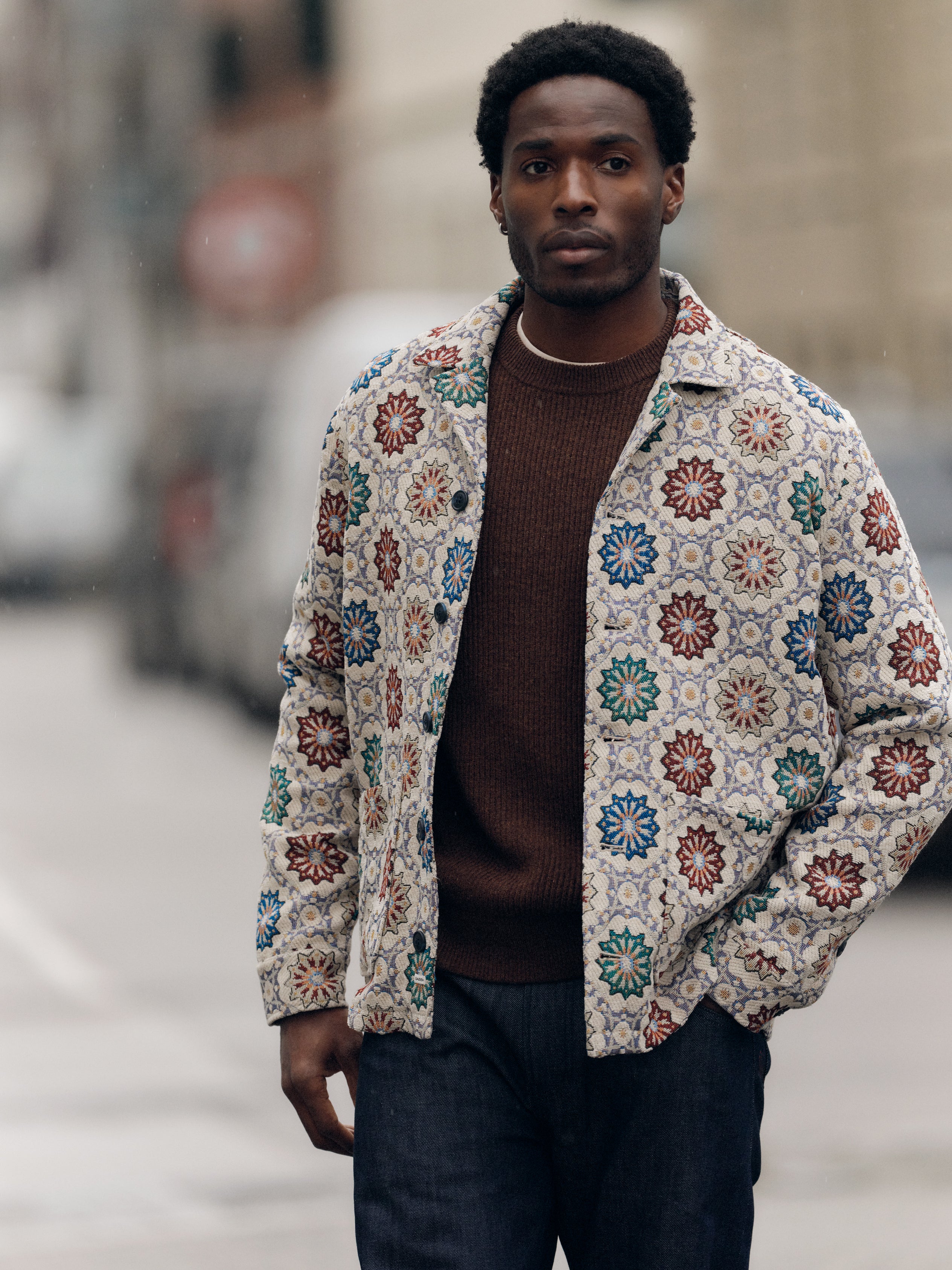 A man stands outdoors wearing the Gabba GABsimone Nava LS - Birch shirt, featuring colorful floral patterns and a resort collar, layered over a brown sweater and dark jeans, with a blurred urban background behind him.