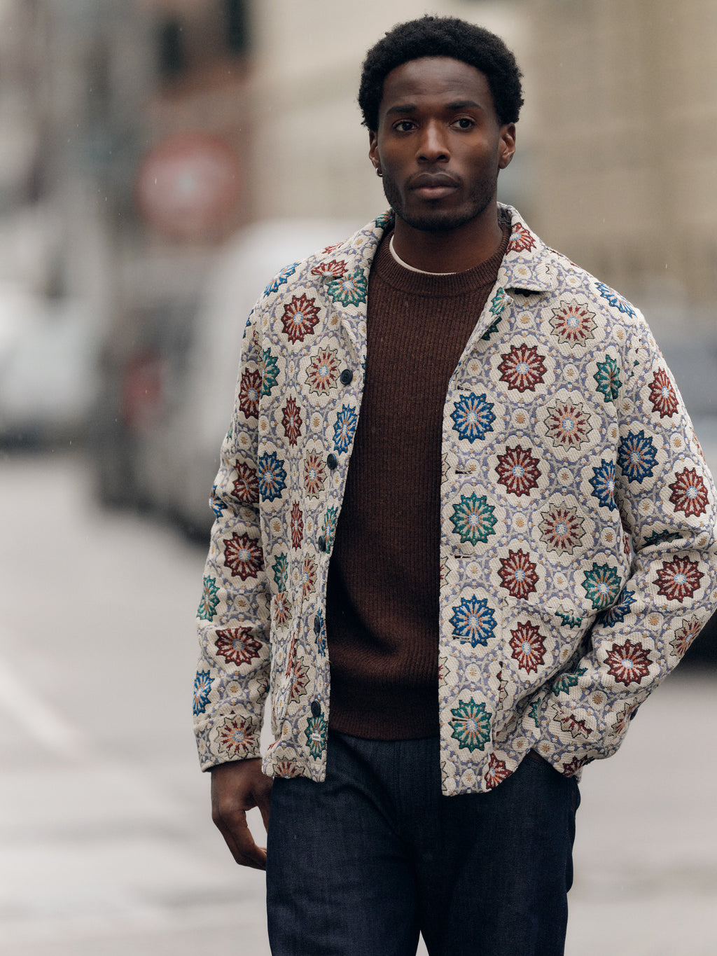 A man stands outdoors wearing the Gabba GABsimone Nava LS - Birch shirt, featuring colorful floral patterns and a resort collar, layered over a brown sweater and dark jeans, with a blurred urban background behind him.