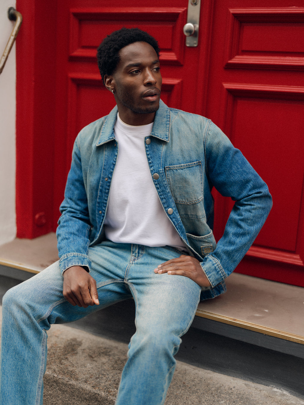 A person wearing a denim jacket in a mid-blue wash, a white shirt, and Gabba GABcarl Bieffe GW0001 Jeans in Mid Blue Denim with a loose tapered fit crafted from 3x1 slub denim sits on outdoor steps in front of a bright red door, looking to the side.