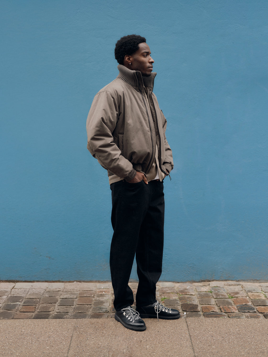 A person in a taupe jacket, Gabba GABsenna Black DS0006 Jeans in black denim with a loose tapered fit, and black sneakers stands on a cobblestone sidewalk against a blue wall, facing left with hands in pockets.