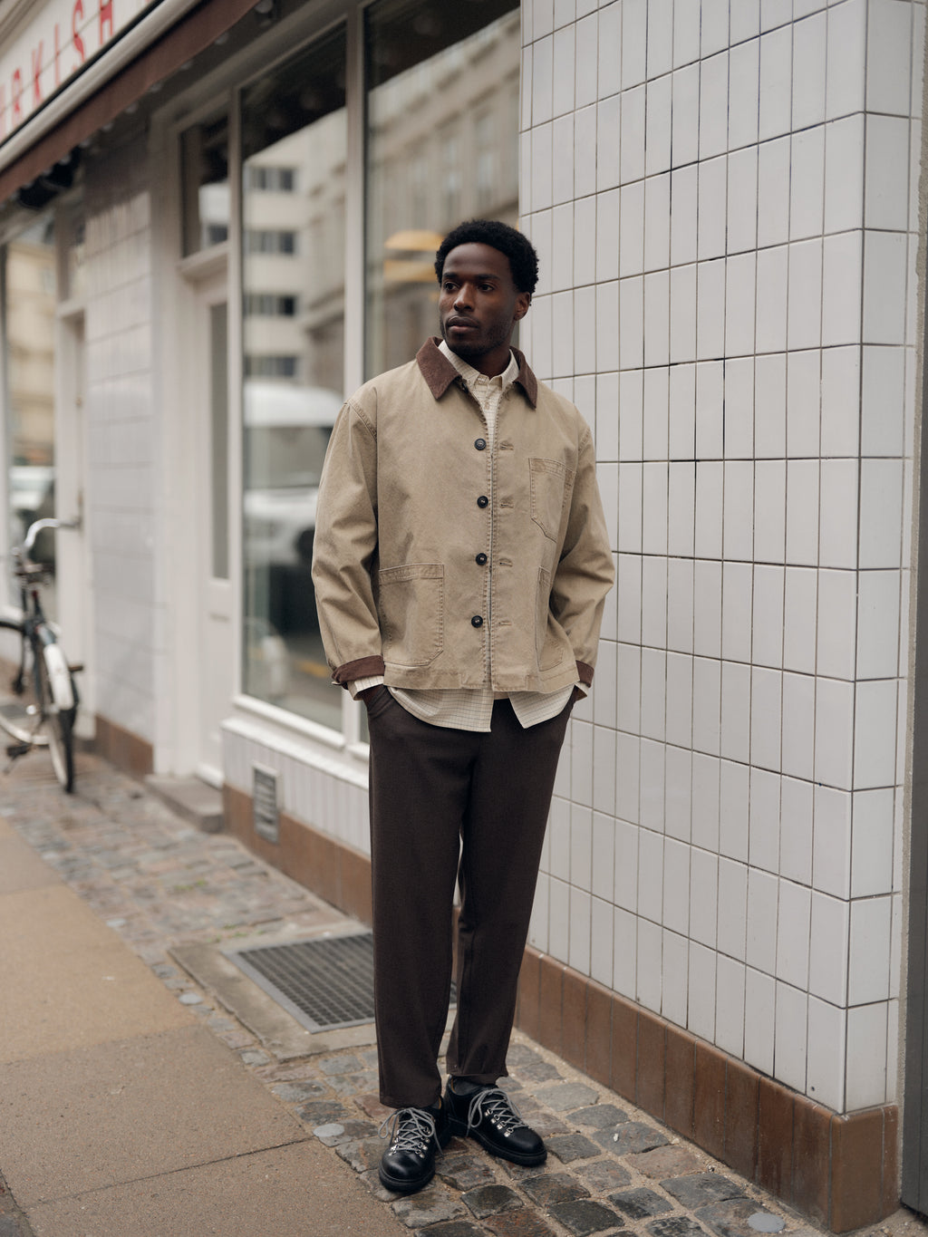 A man wearing a beige jacket, black shoes, and Gabba’s GABdavid Rubin Pant in Brown Slate stands on a city sidewalk near a tiled building wall, with a bicycle and windows in the background.