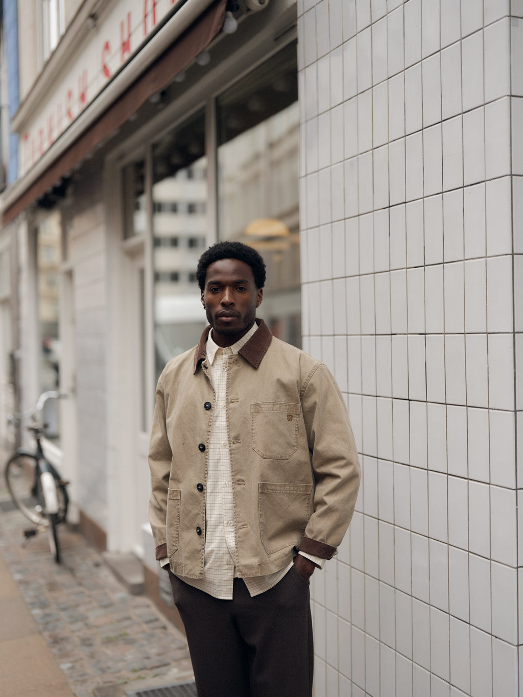 A man wearing a beige jacket and dark pants stands on a city sidewalk near a tiled building, his look complemented by the Gabba GABjens Over Indie LS Shirt in Lt. Sand—a cotton-blend oversized shirt—with a bicycle parked nearby.