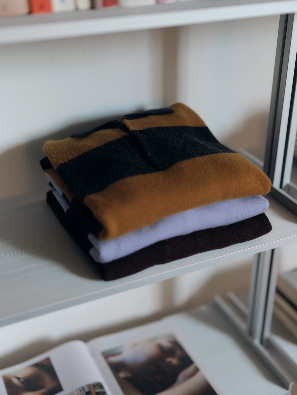 Three folded sweaters—one GABworren Polo Stripes Aico LS Knit in yellow stripes from Gabba, one solid lavender, and one solid dark brown—are stacked on a white shelf. A book with photos lies open on the lower shelf, adding a cozy touch to this Gabba display.