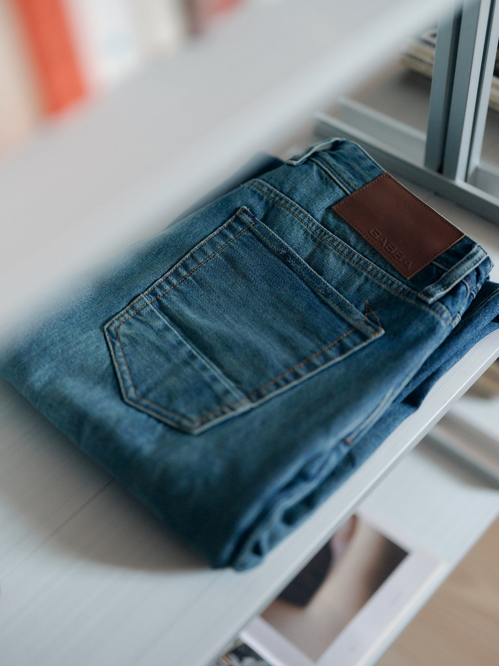 A pair of Gabba GABcarl Bieffe GW0001 Jeans in mid blue denim, featuring a brown leather patch on the waistband and crafted from rigid 3x1 denim, rests folded on a light-colored shelf.