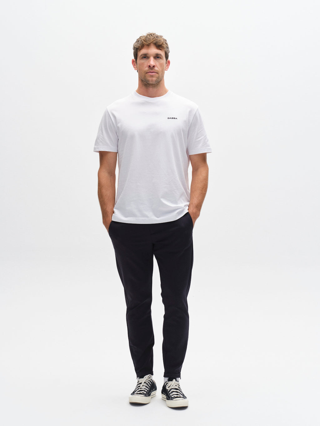 A man faces forward against a plain white background, wearing a white t-shirt, GABBA Pisa Small Dot - Navy pants in stretch cotton-blend, and black sneakers with white soles. His metro style is relaxed, with his arms at his sides.