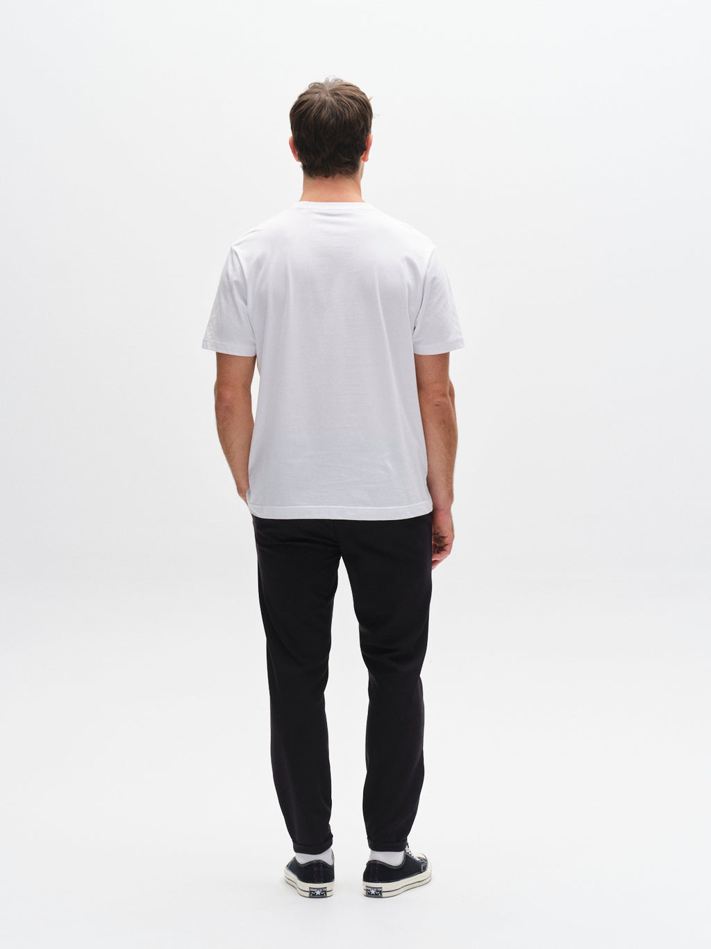 A man faces away from the camera in a plain white t-shirt, GABBA Pisa Small Dot - Black pants, and black slip-on shoes, showcasing modern metro style against a white background.