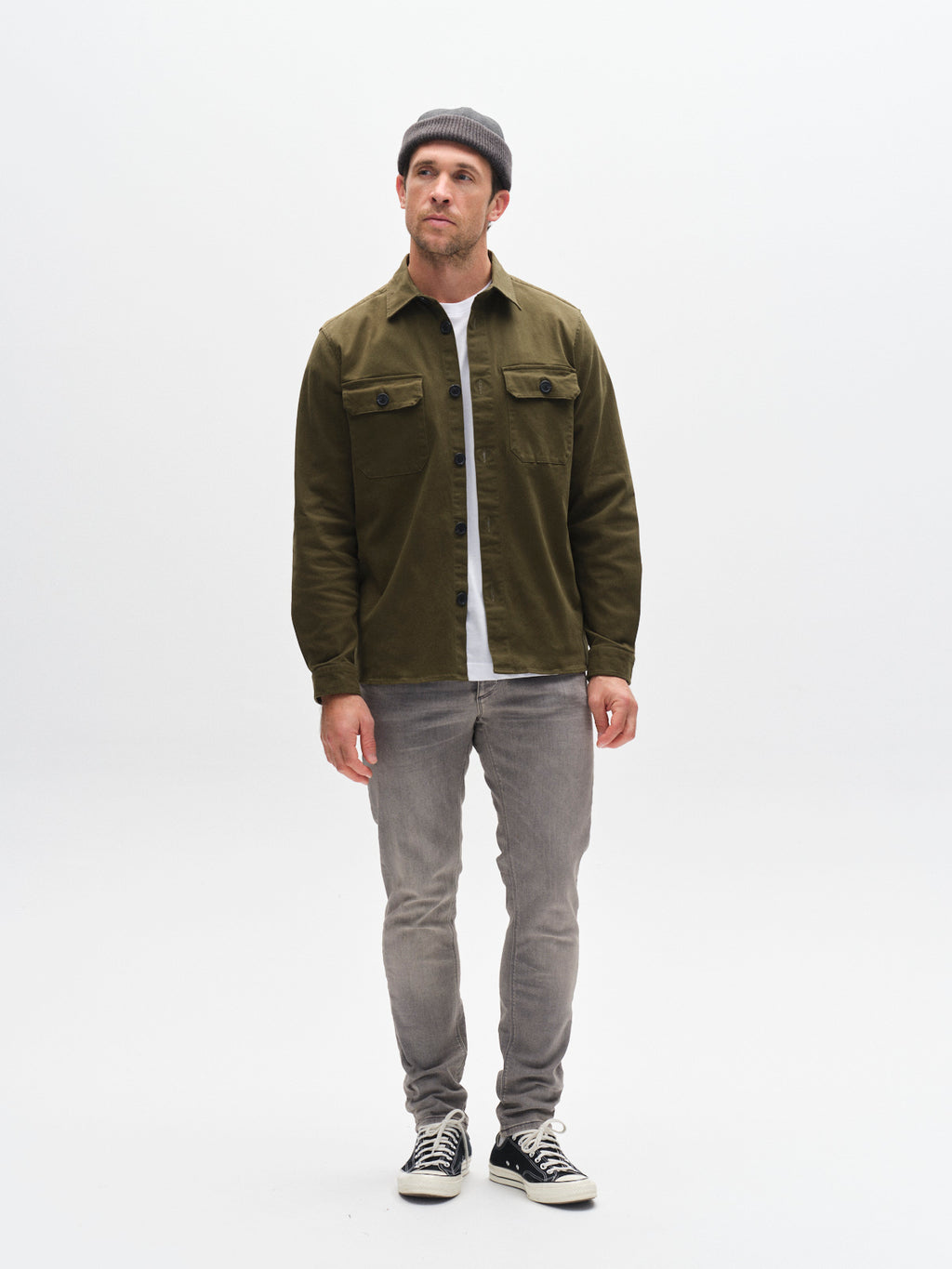 A man in GABBA's GABrey K3454 - Denim wash jeans stands against a white background, wearing a gray beanie, olive jacket, white tee, and black sneakers, looking slightly upward with a neutral expression for a relaxed vibe.