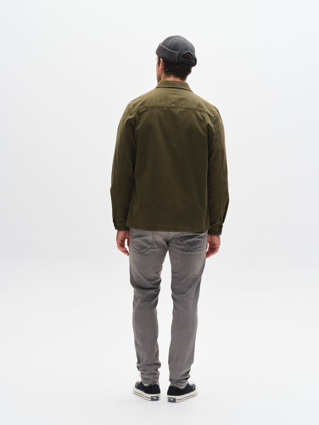 A person stands facing away from the camera against a white background, wearing GABBA's GABrey K3454 - Denim wash jeans, a gray beanie, olive green jacket, and black shoes.