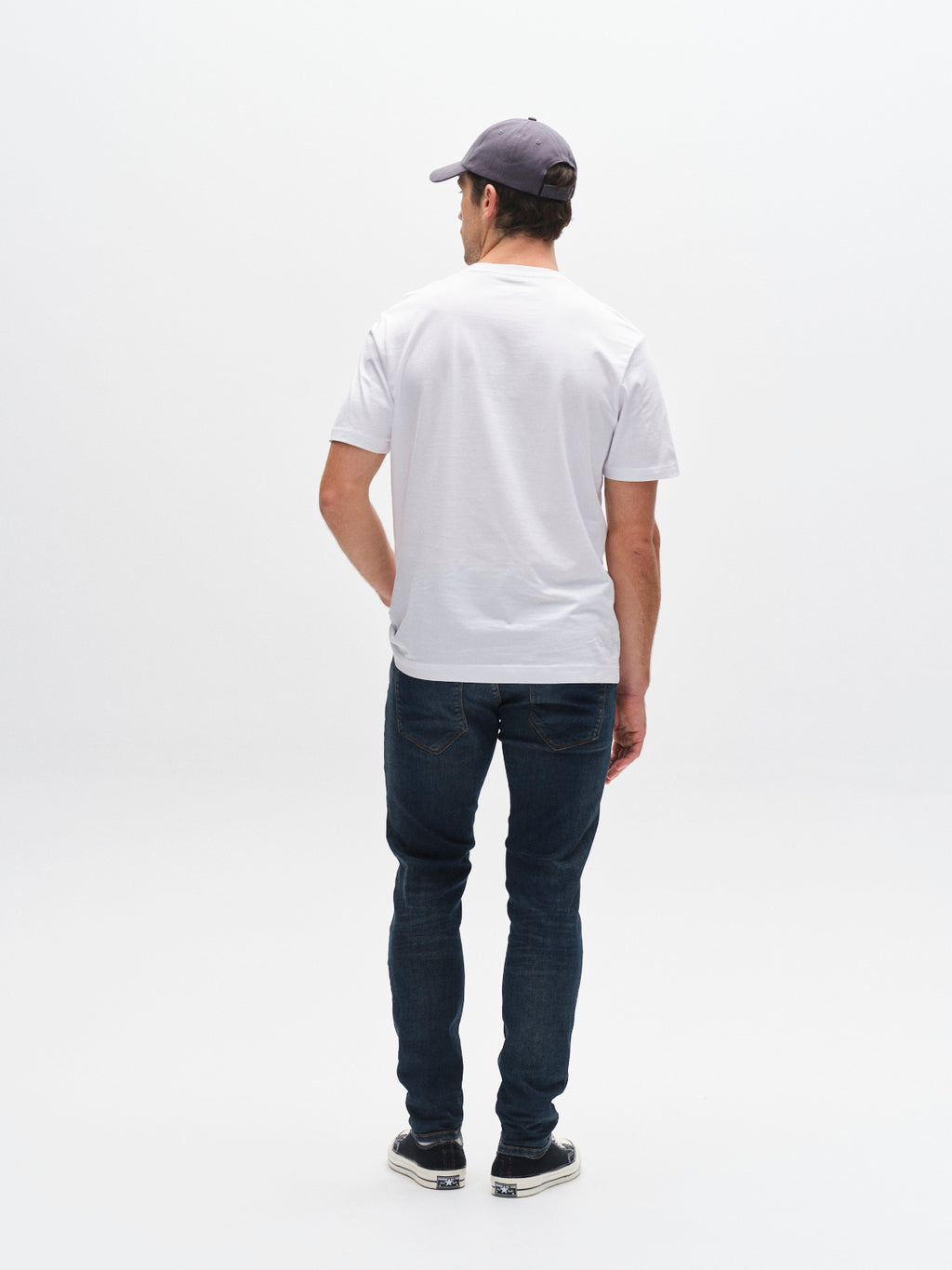 A man wearing a gray cap, white t-shirt, GABBA GABrey K3606 Mid Blue Jeans in mid blue denim, and sneakers stands facing away from the camera against a plain white background for a laid-back look.