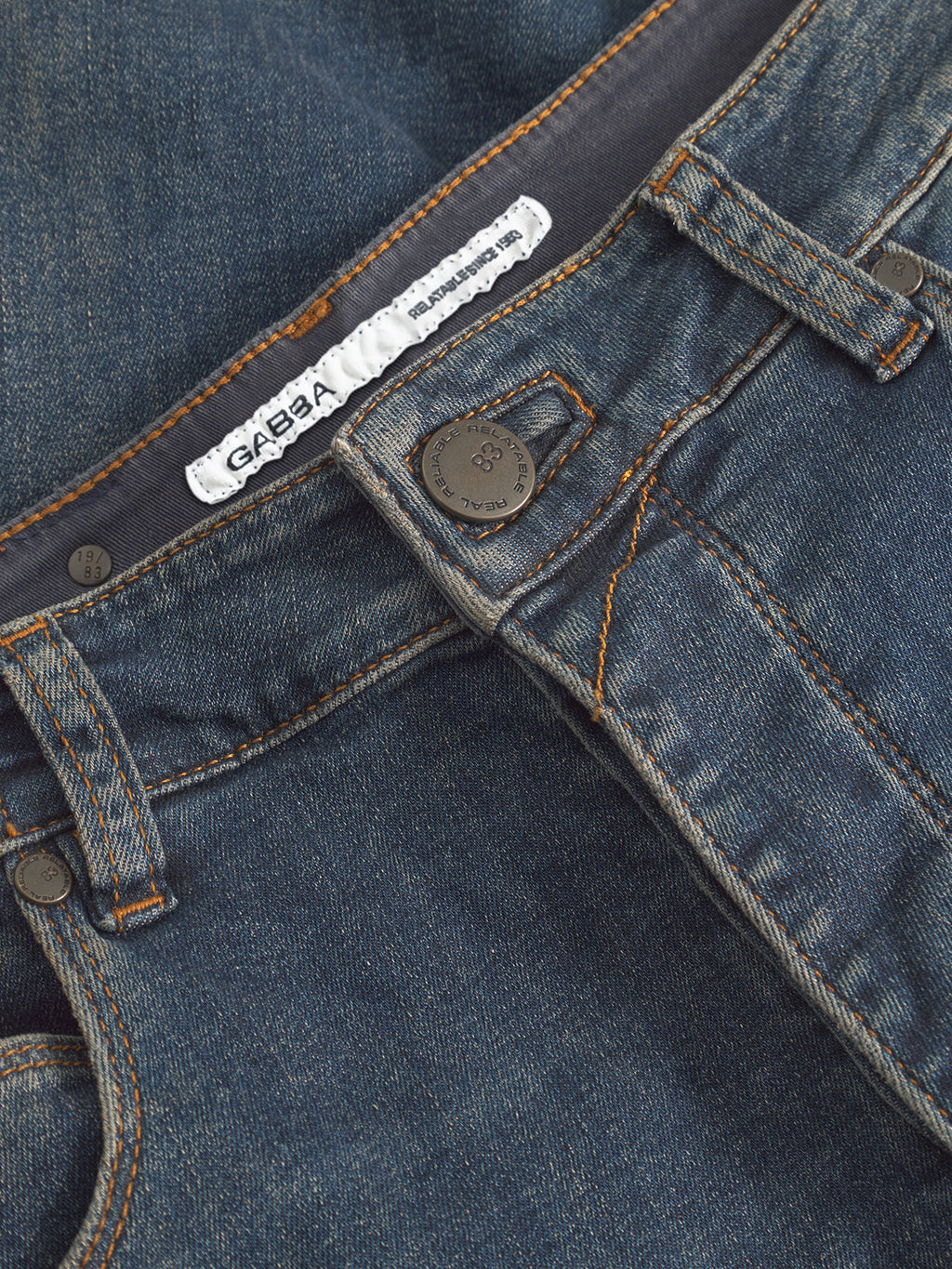 Close-up of GABBA GABrey K3606 Mid Blue Jeans in mid blue denim, showing waistband, belt loops, button, zipper, and orange stitching. The inside white label displays the brand name for a relaxed look.