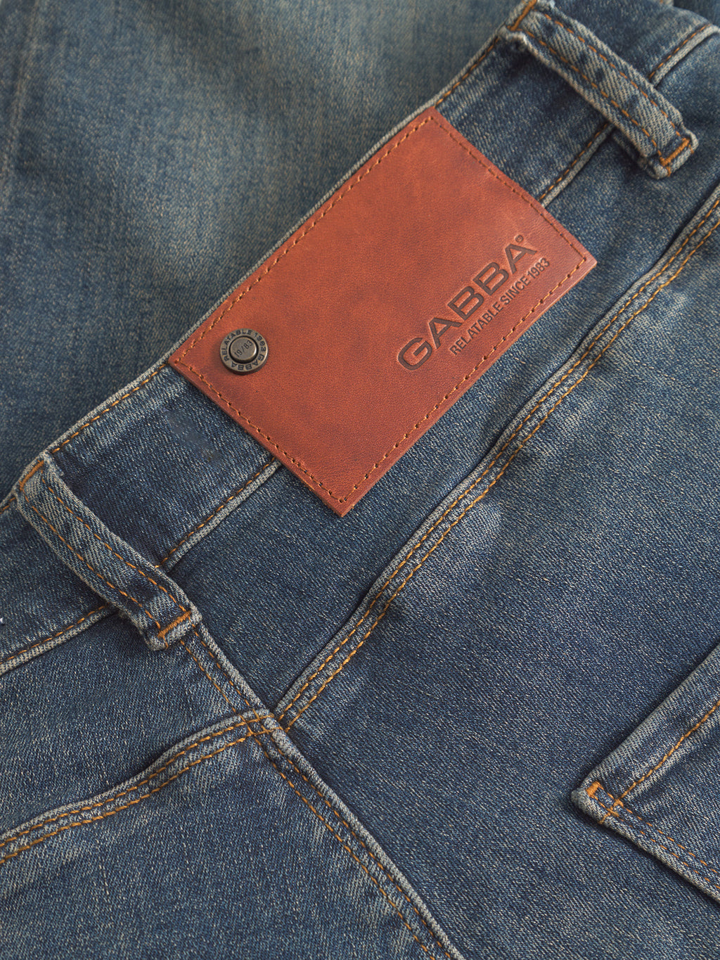 Close-up of GABBA GABrey K3606 Mid Blue Jeans in mid blue denim, featuring a brown leather patch label on the waistband, metal button, visible belt loops, pockets, and orange stitching for a relaxed style.