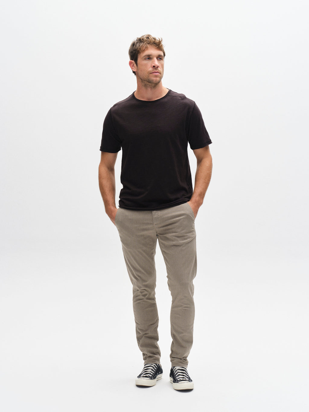 A man stands against a white background, wearing a black t-shirt, GABBA GABpaul K3280 Dale Chino in Humus with French pockets, and black-and-white sneakers. His hands are in his pockets as he looks slightly to the side.