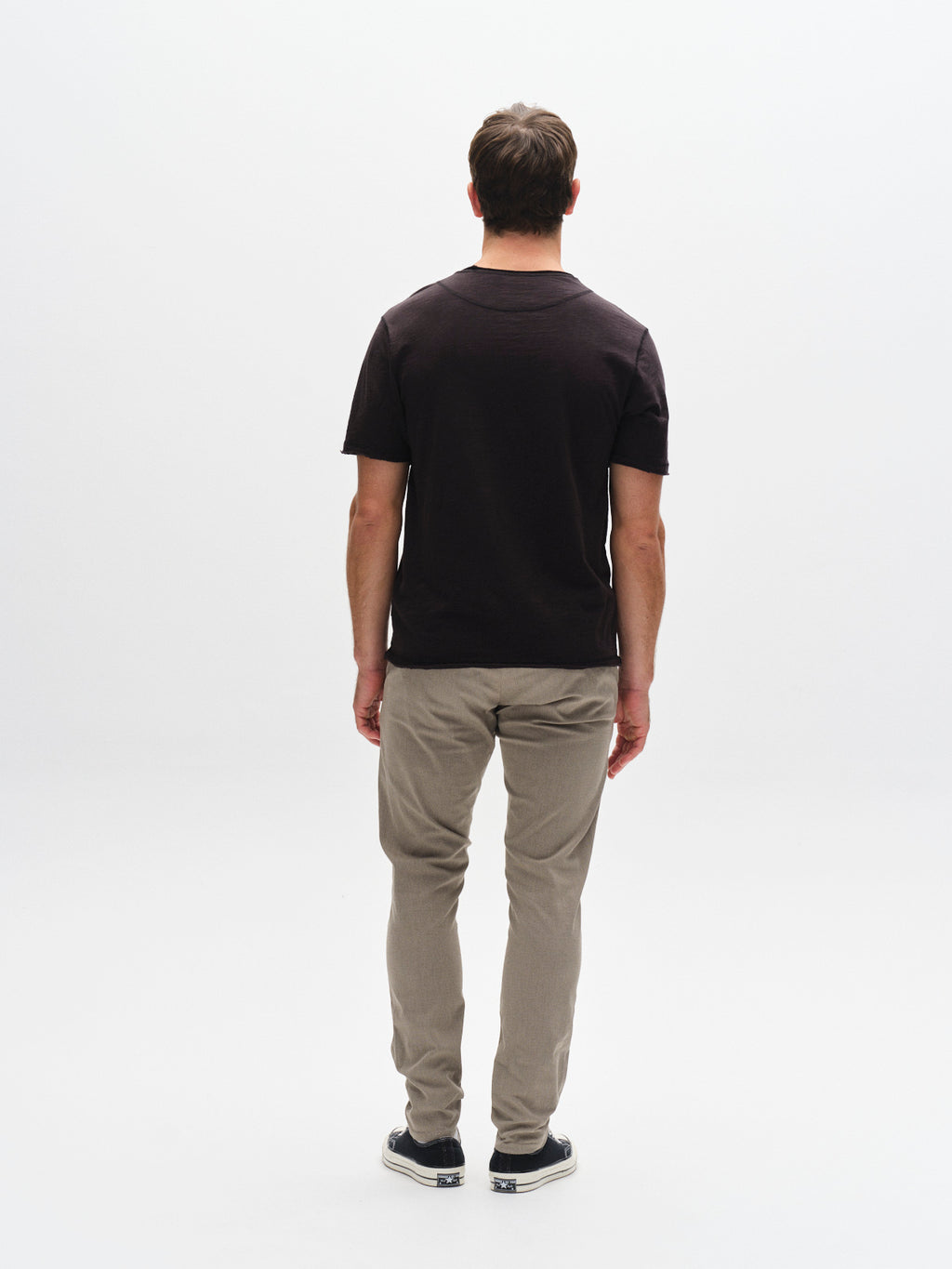 A man stands facing away from the camera, wearing a black t-shirt, black shoes, and GABBA GABpaul K3280 Dale Chino - Humus slim-fit pants with French pockets, set against a plain white background.