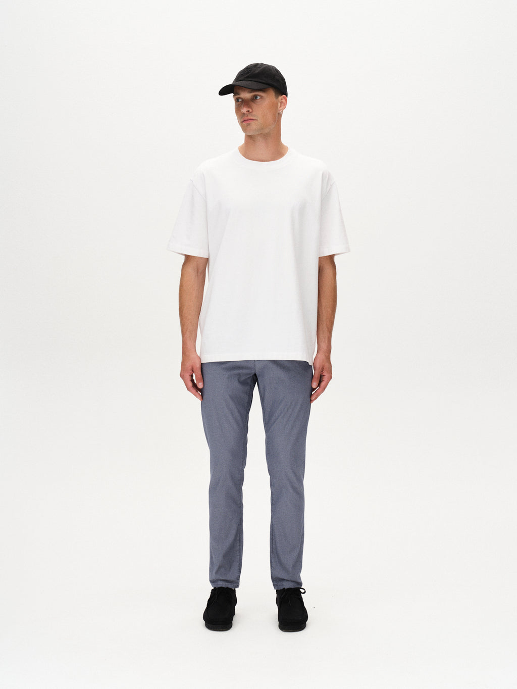 A man wears a black cap, white T-shirt, Gabba Paul K3280 Dale Chino in blue, and black shoes, standing against a plain white background with his arms relaxed at his sides and looking off to the side.