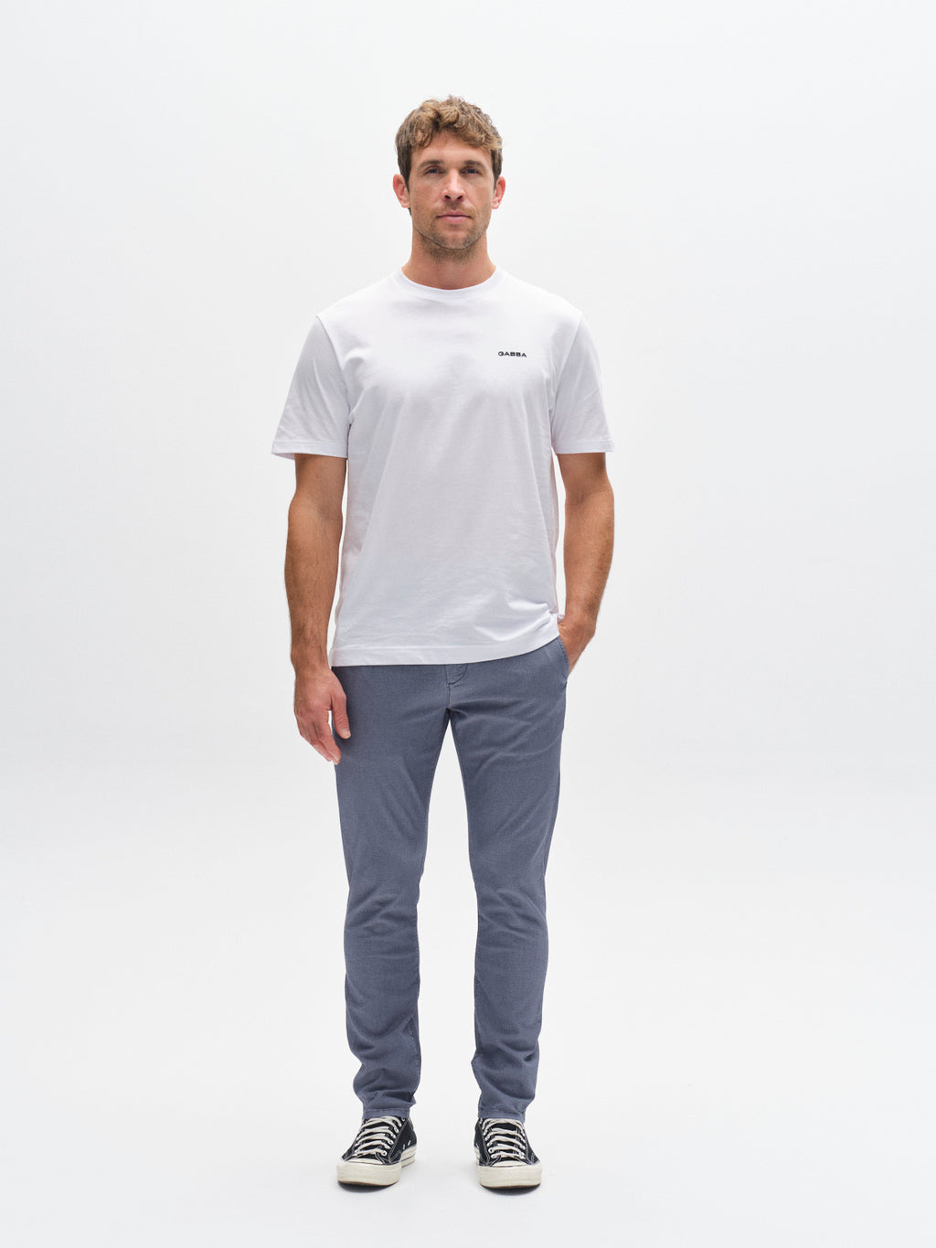 A man stands facing forward against a white background, wearing a white t-shirt and Gabba GABpaul K3280 Dale Chino - Blue, paired with black sneakers with white laces. He has one hand in his pocket and a neutral expression.
