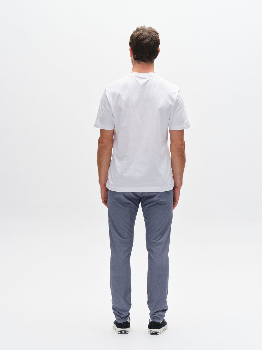 A man stands with his back to the camera in a white studio, wearing a white short-sleeve t-shirt, Gabba GABpaul K3280 Dale Chino in blue, and black shoes. The background is entirely white.