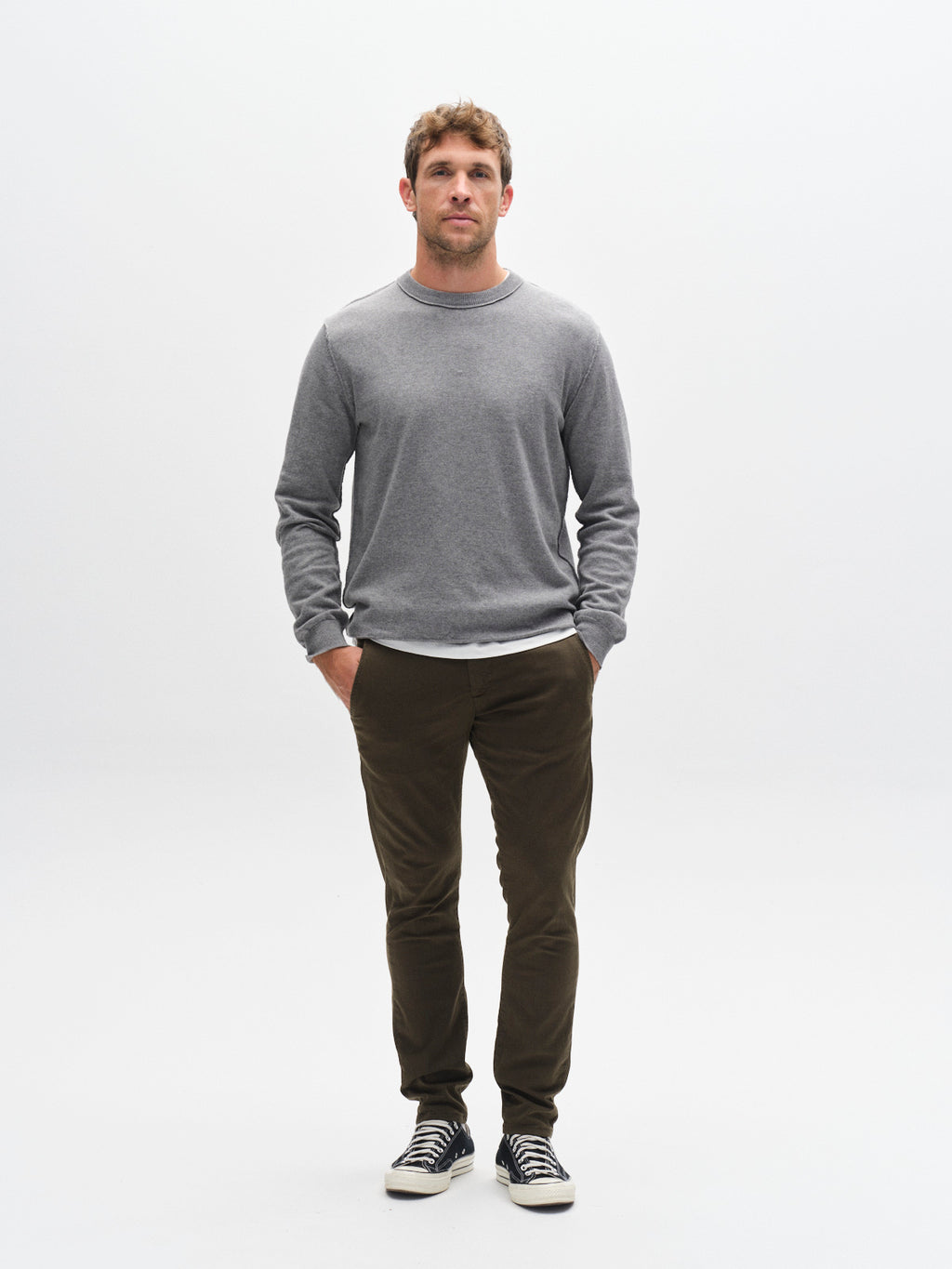 A man with short brown hair stands facing forward, hands in the French pockets of his GABBA GABpaul K3280 Dale Chino - Army pants. He wears a gray sweater and black sneakers with white soles, set against a plain white background.