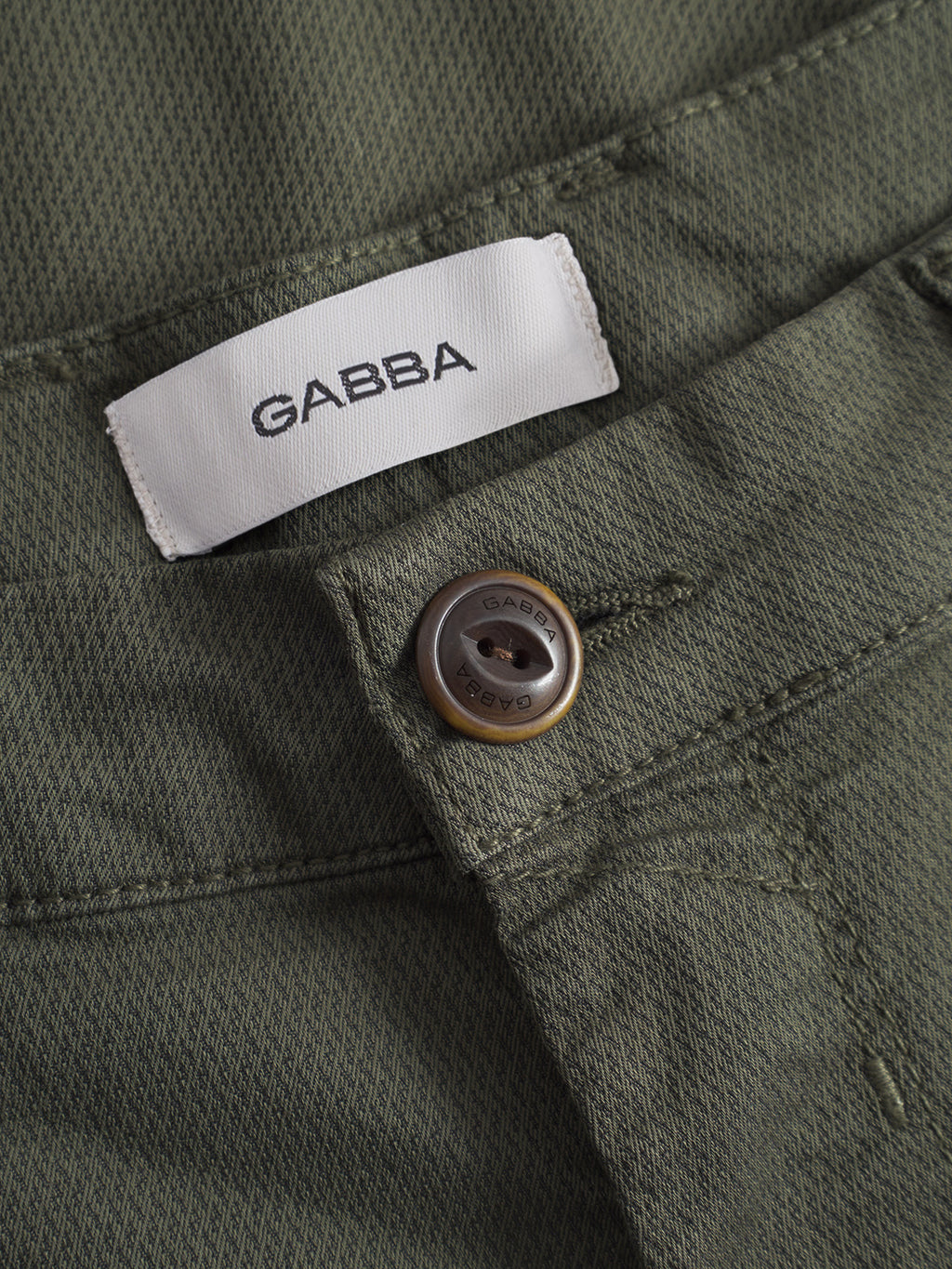 Close-up of GABBA's GABpaul K3280 Dale Chino - Army: green textured slim-fit pants with French pockets, white GABBA label, and a dark GABBA-engraved button near the waistband.