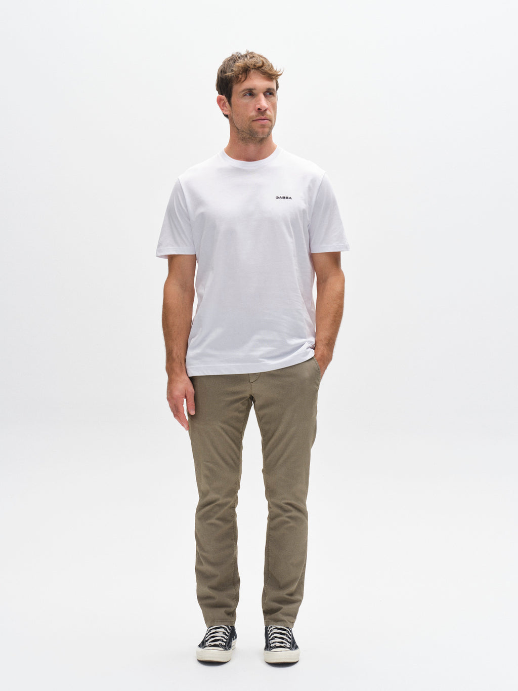 A man stands against a plain white background wearing the Gabba GABpaul K3280 Dale Chino in olive green, paired with a white t-shirt and black-and-white sneakers, looking slightly to the side with his hands relaxed at his sides.