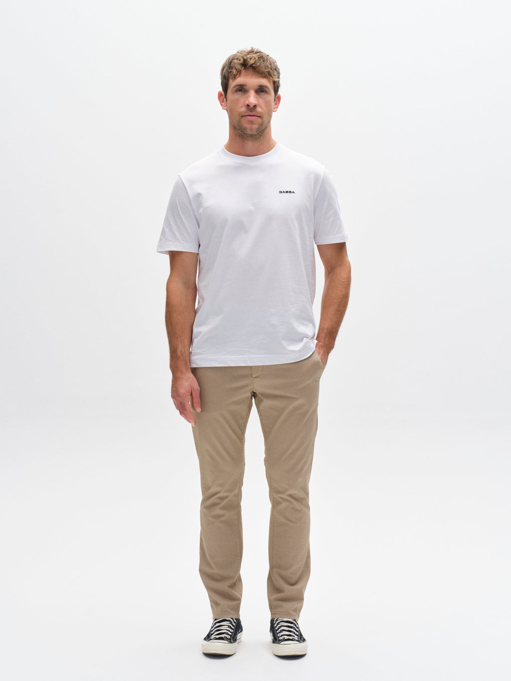 A man stands facing forward against a white background, wearing a white T-shirt, Gabba GABpaul K3280 Dale Chino - Sand pants with French pockets, and black-and-white sneakers. His arms are relaxed at his sides.